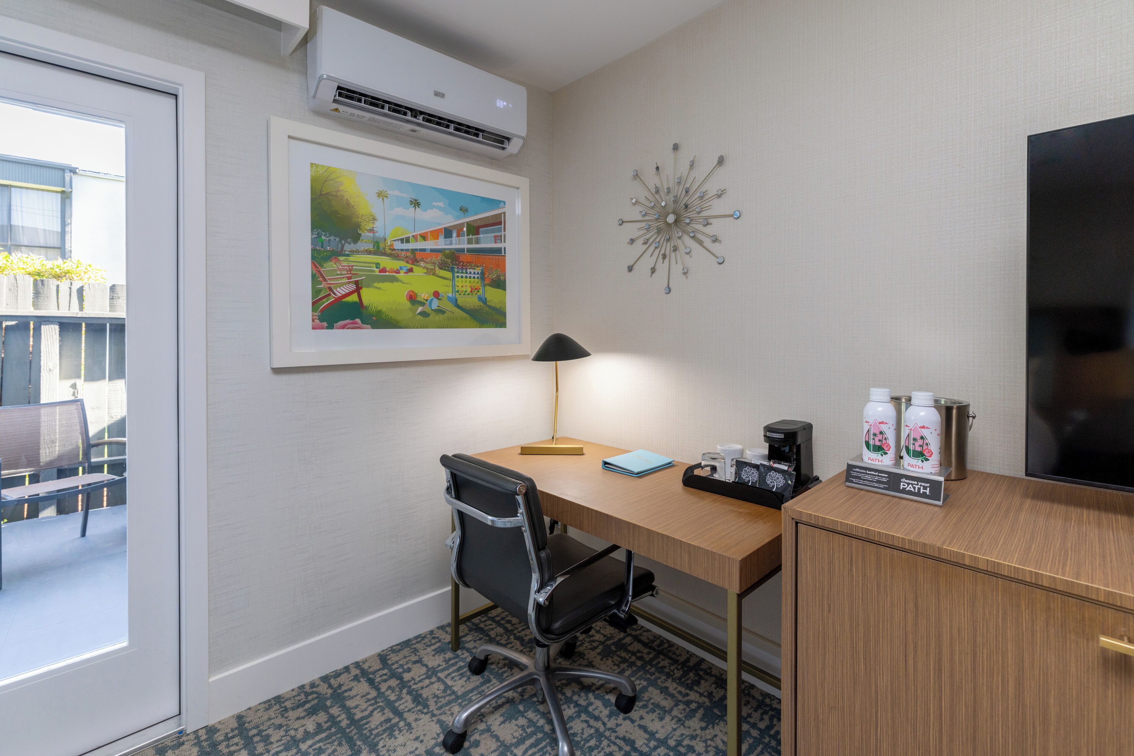 A hotel room with a desk, chair, air conditioning unit, artwork, decorative wall piece, and a television.