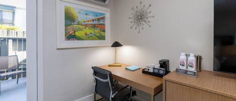 A hotel room with a desk, chair, air conditioning unit, artwork, decorative wall piece, and a television.