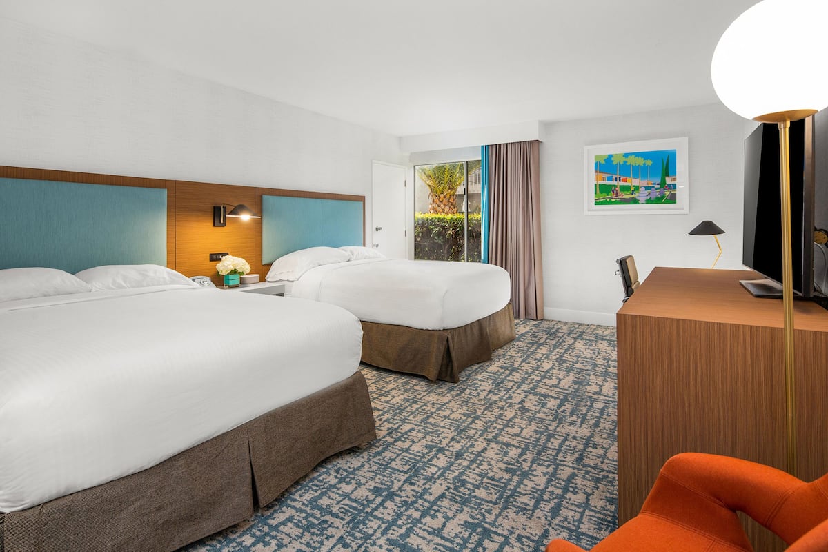 A hotel room with two beds, a desk, a chair, and a large window with a view of greenery.