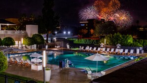 A pool area with lounge chairs and umbrellas, surrounded by greenery and a building with illuminated signage. - The Anaheim Hotel (Anaheim)