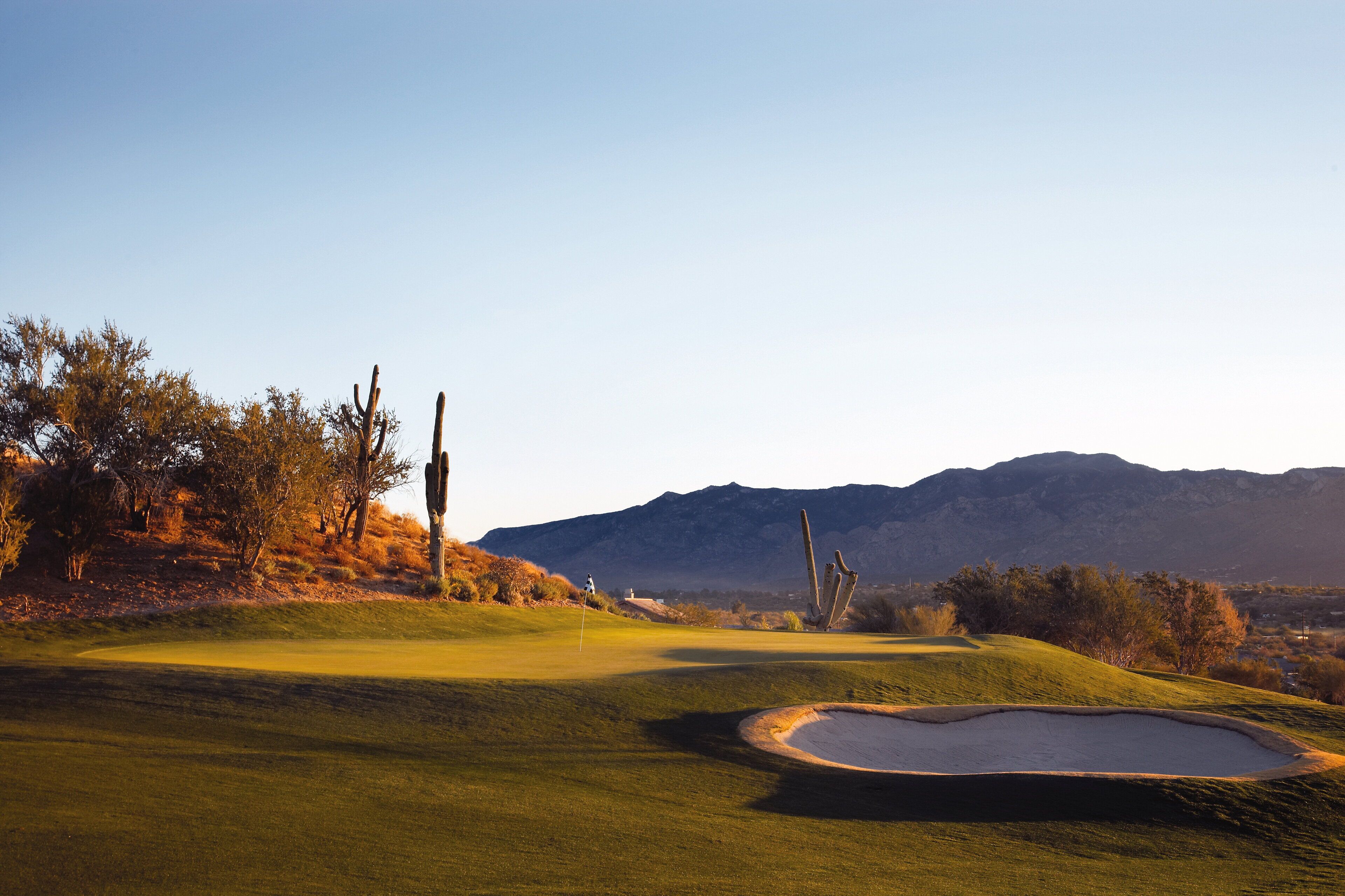 Un campo de golf con cielo despejado, montañas al fondo y una zona verde bien cuidada con un búnker de arena.