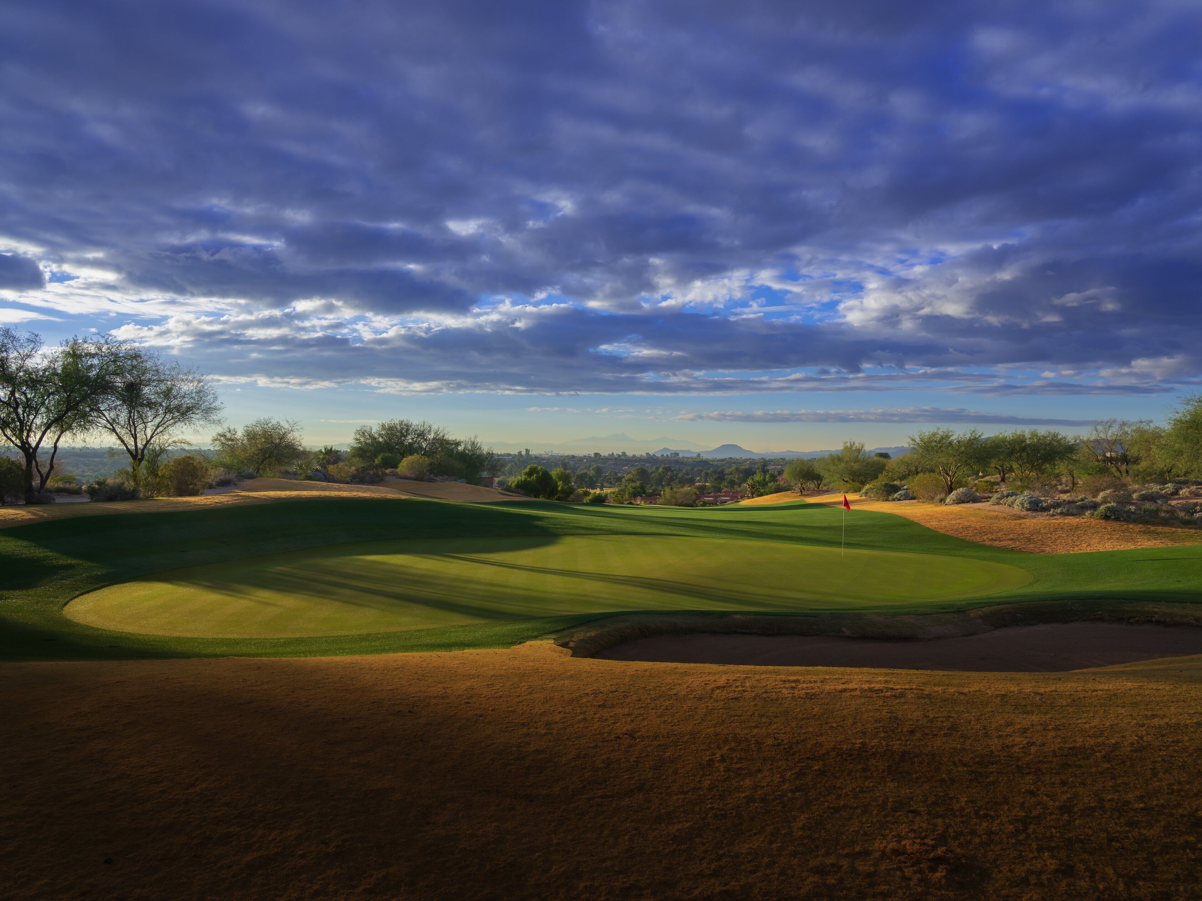 A golf course with a flag, trees, and a distant cityscape under a cloudy sky.