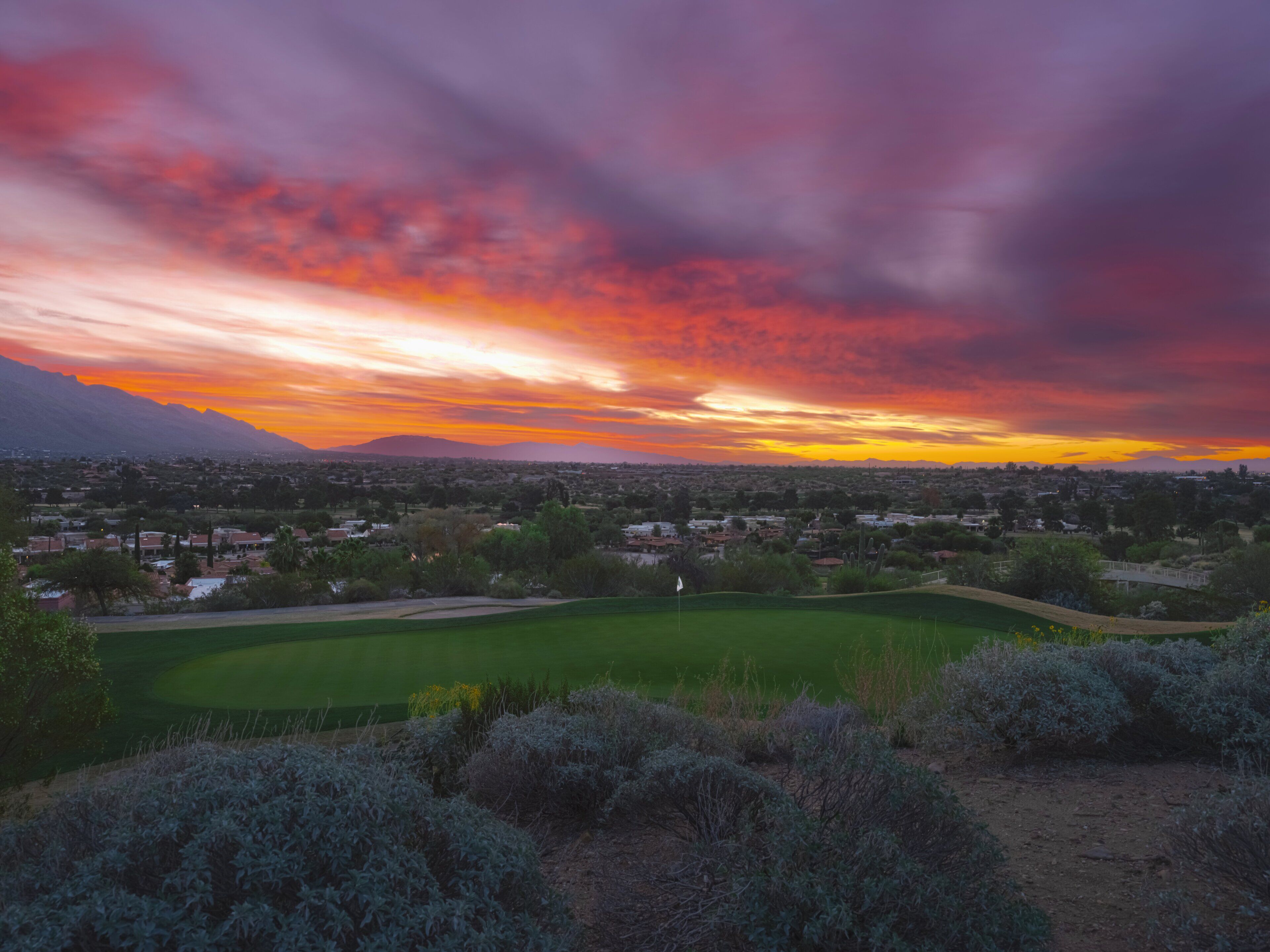 A golf course with a flag, surrounded by shrubbery and a view of a cityscape during sunset.