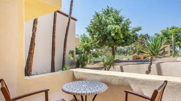A patio with a round mosaic table and two wooden chairs, overlooking a landscaped garden with trees and a cactus.