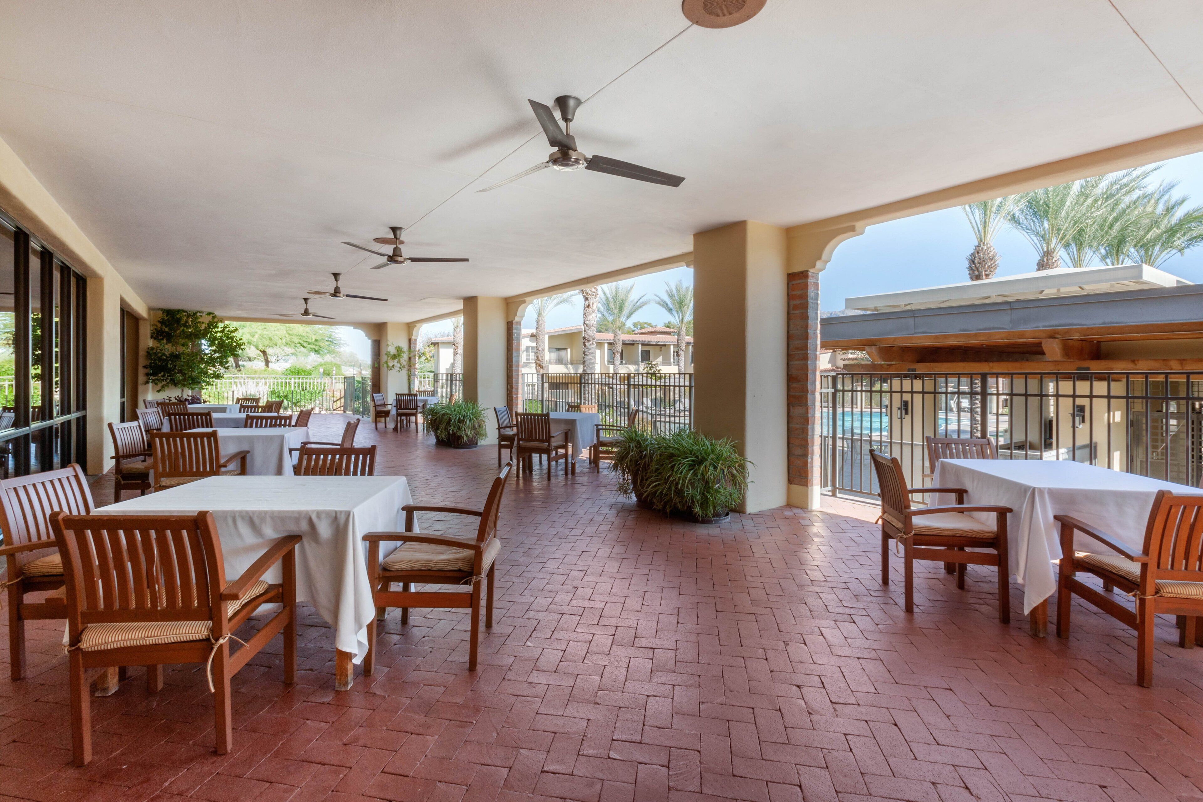 A covered outdoor dining area with brick flooring, wooden tables and chairs, ceiling fans, and a view of palm trees and buildings in the background.