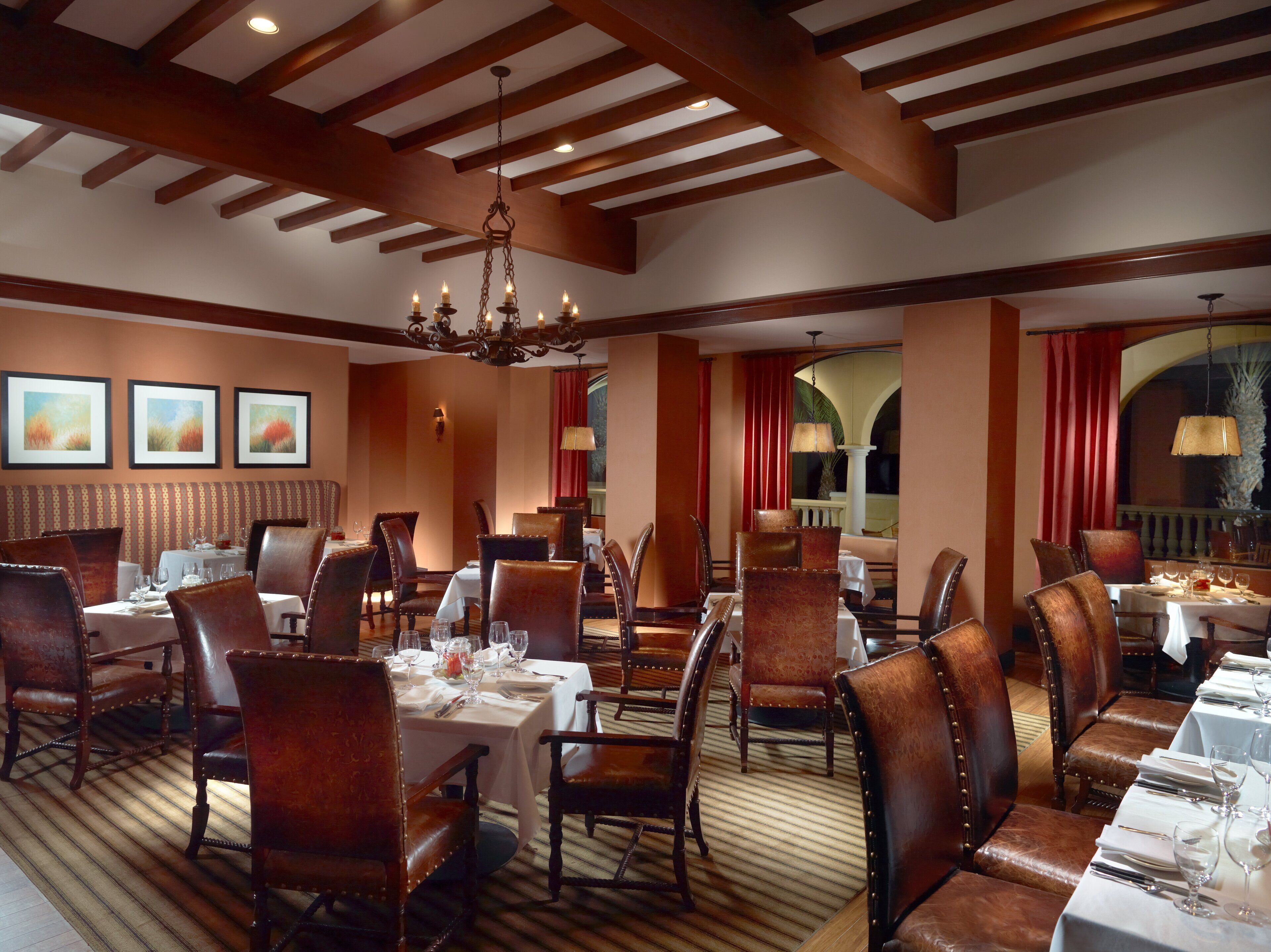 A formal dining room with multiple tables and chairs, wooden beams on the ceiling, and framed artwork on the walls.