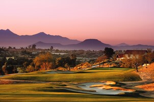 Un campo de golf con un club house, búnkeres de arena y montañas al fondo.
