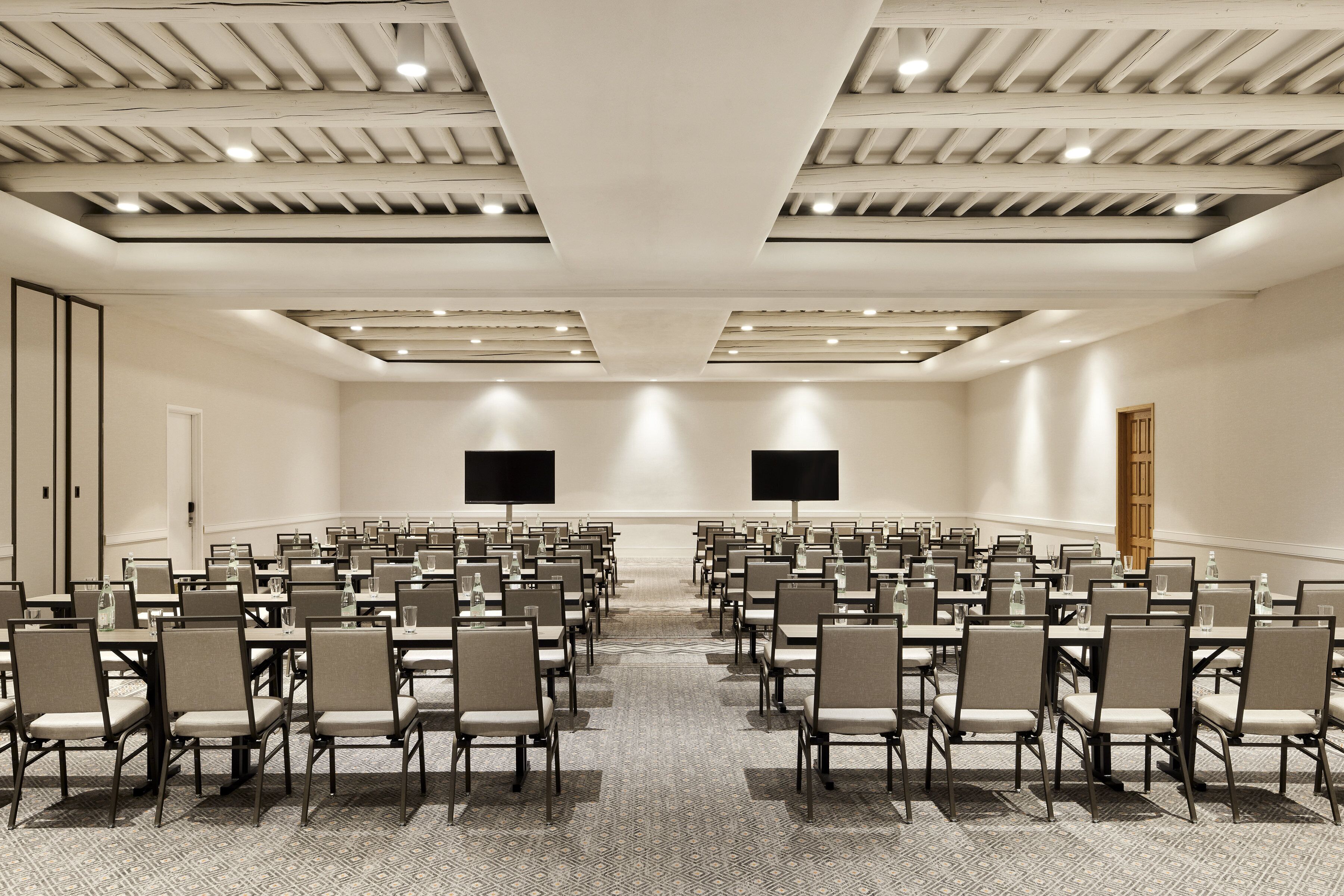 A conference room with rows of chairs and tables, a high ceiling with recessed lighting, and a large screen at the front.