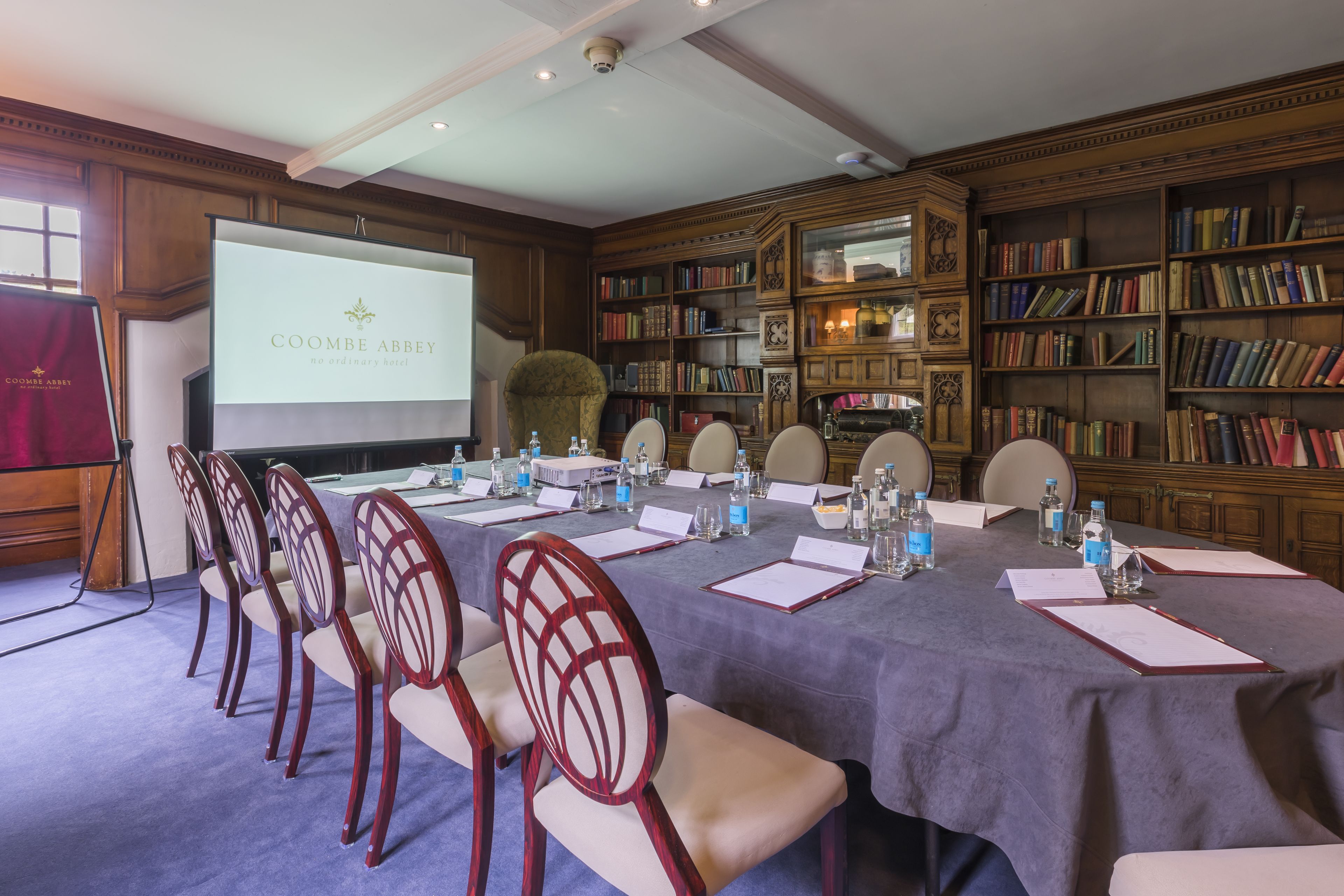 Un salón de conferencias con una larga mesa, sillas, una pantalla de proyector y una estantería.