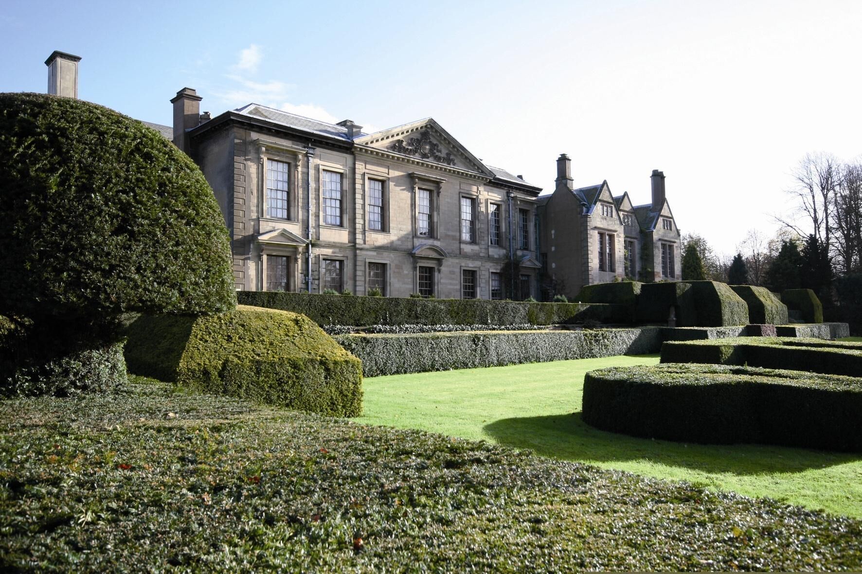 A large, historic building with symmetrical architecture, a well-manicured garden with hedges and a lawn, and a clear sky.