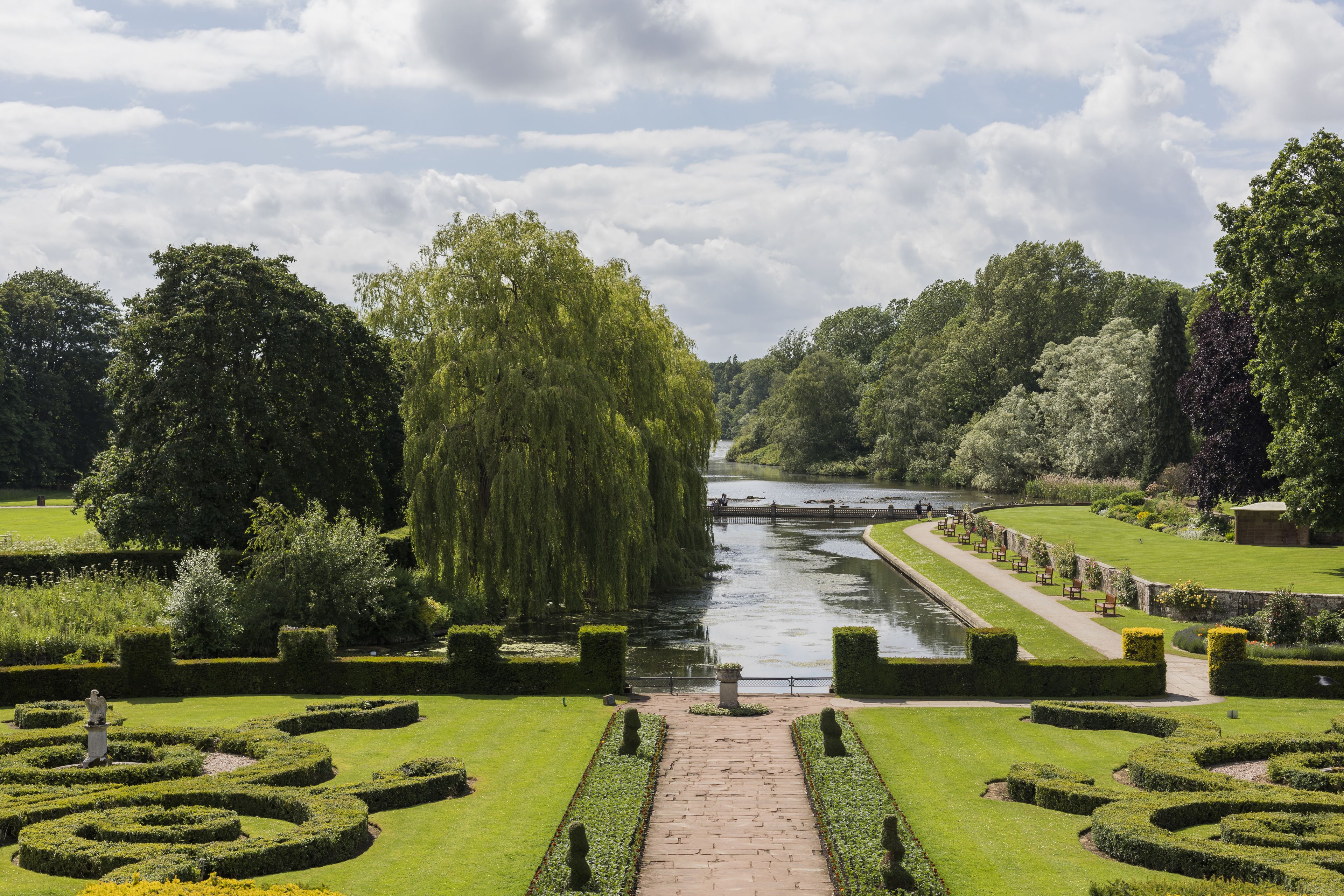 A formal garden with a central pathway leading to a body of water, surrounded by neatly trimmed hedges and trees.