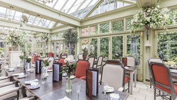 A conservatory with tables set for dining, surrounded by chairs, and adorned with floral arrangements.