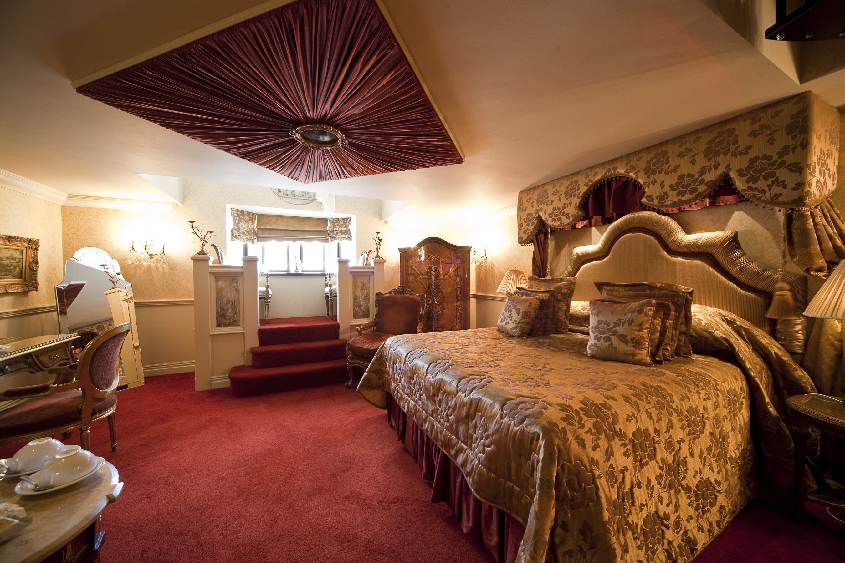 A spacious bedroom with a large bed, a desk, and a chair. There is a staircase leading to an upper level.