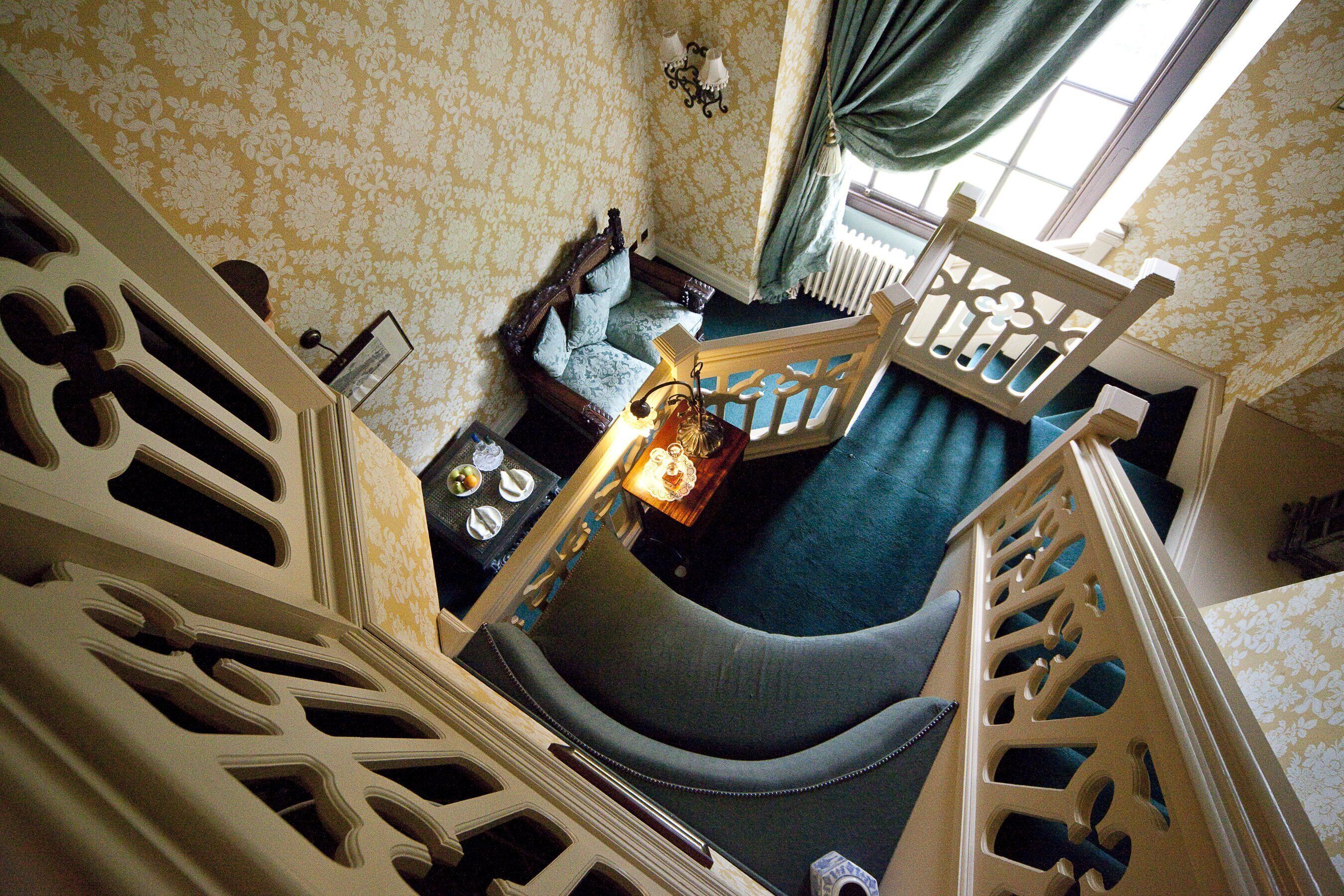 A room with a staircase, a sofa, a table with a lamp, and a window with curtains.
