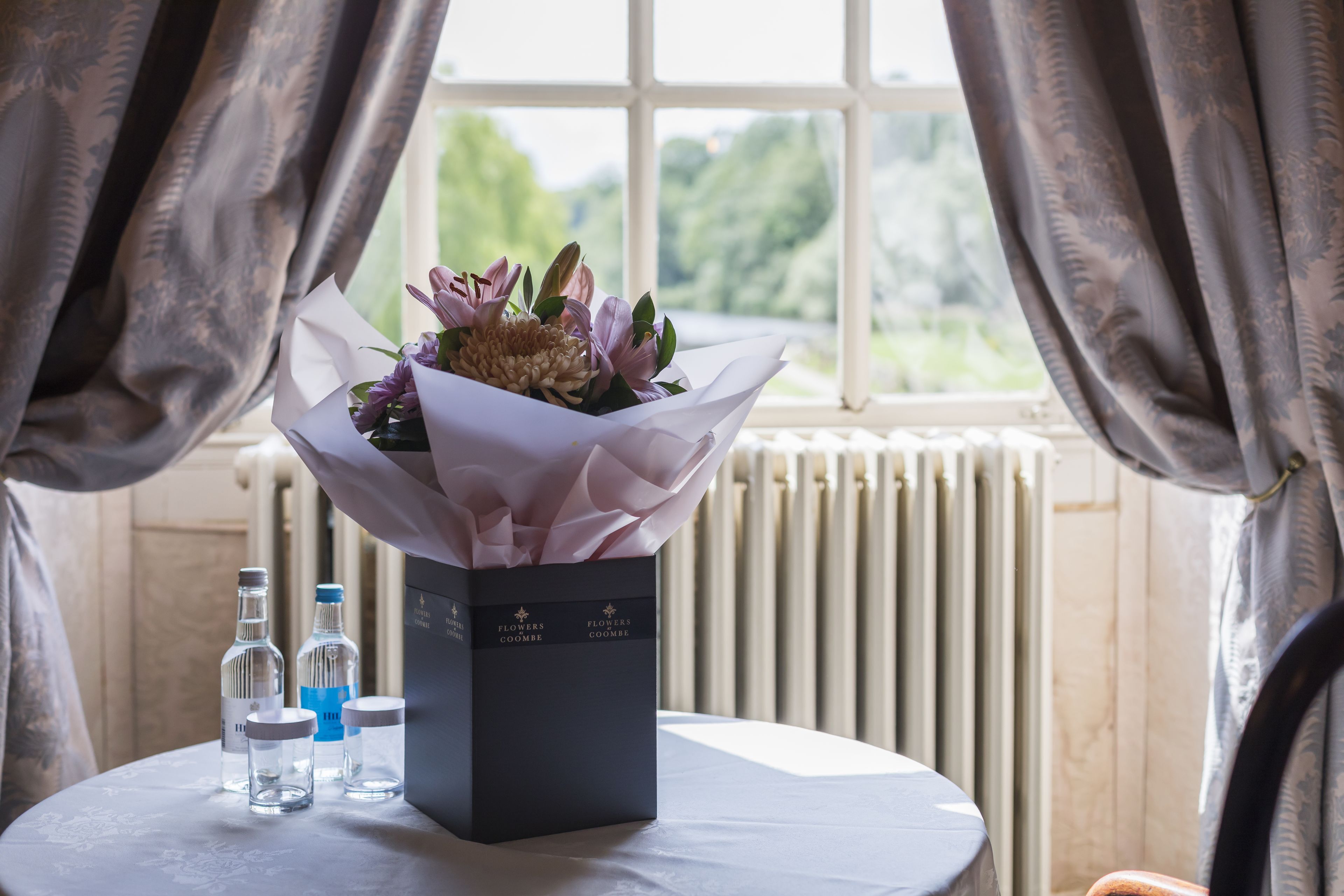 A table with a bouquet of flowers in a box, two bottles, and a radiator in the background.