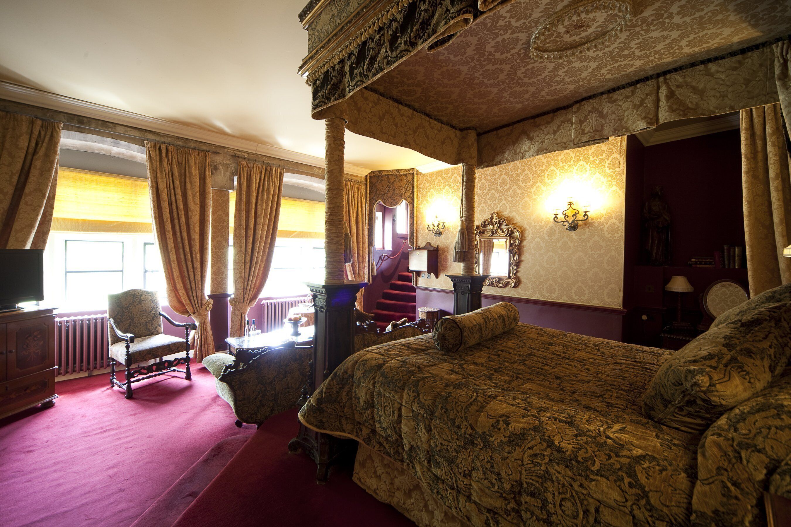 Una camera da letto tradizionale con un letto a baldacchino, una zona salotto con una poltrona e un tavolo, e un'ampia finestra con tende.