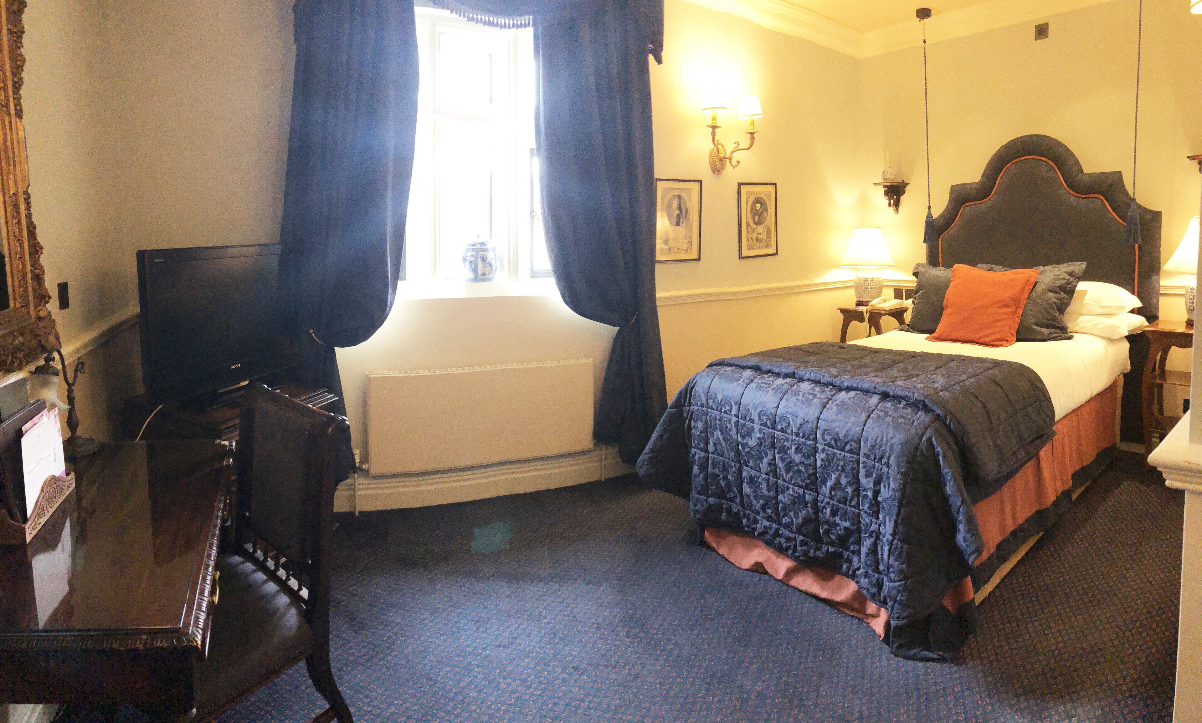 Habitación de hotel con cama, escritorio, silla, ventana con cortinas, radiador y lámparas de pared.
