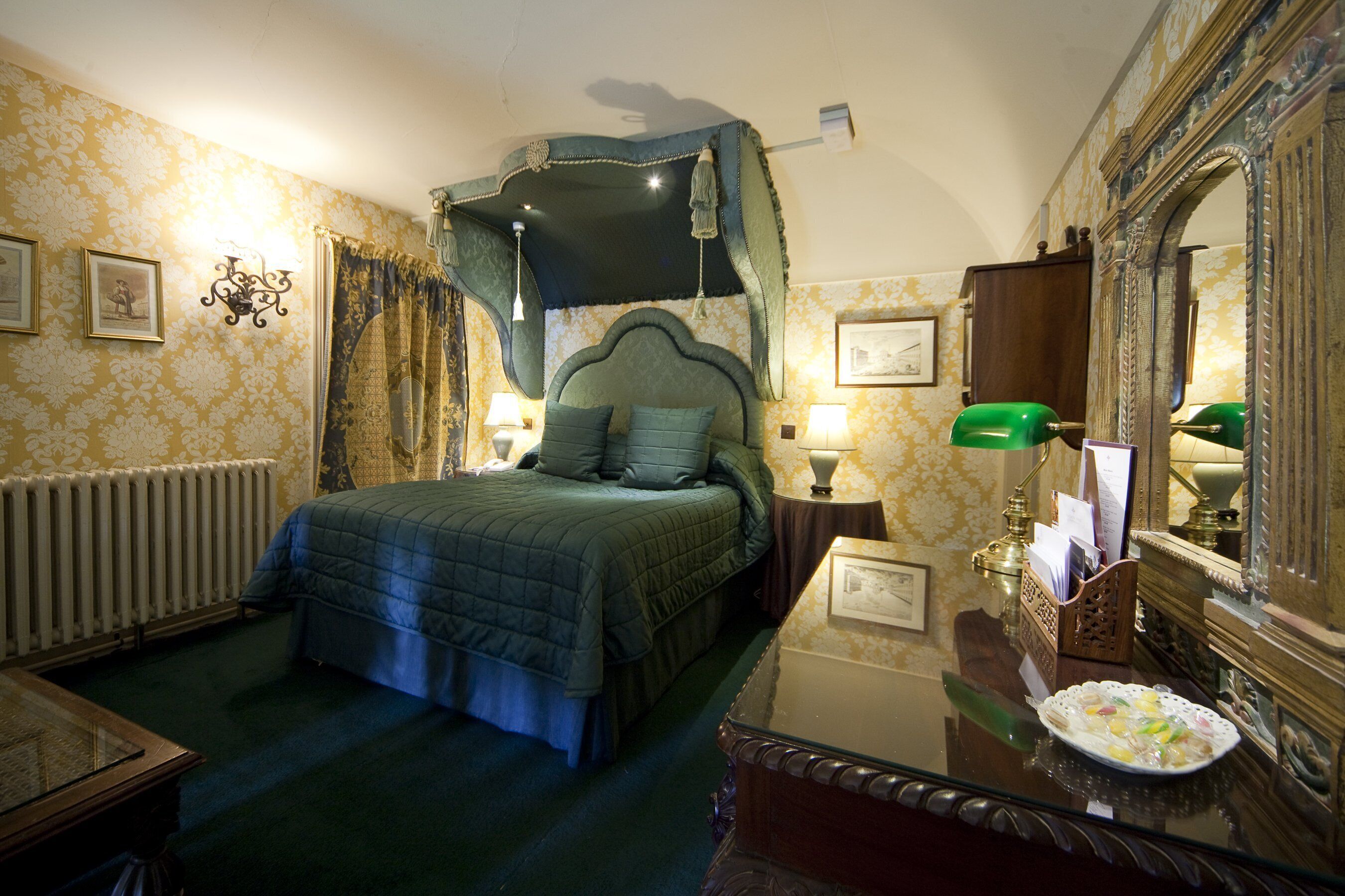 A bedroom with a four-poster bed, a nightstand with a lamp, a dresser, and a mirror.