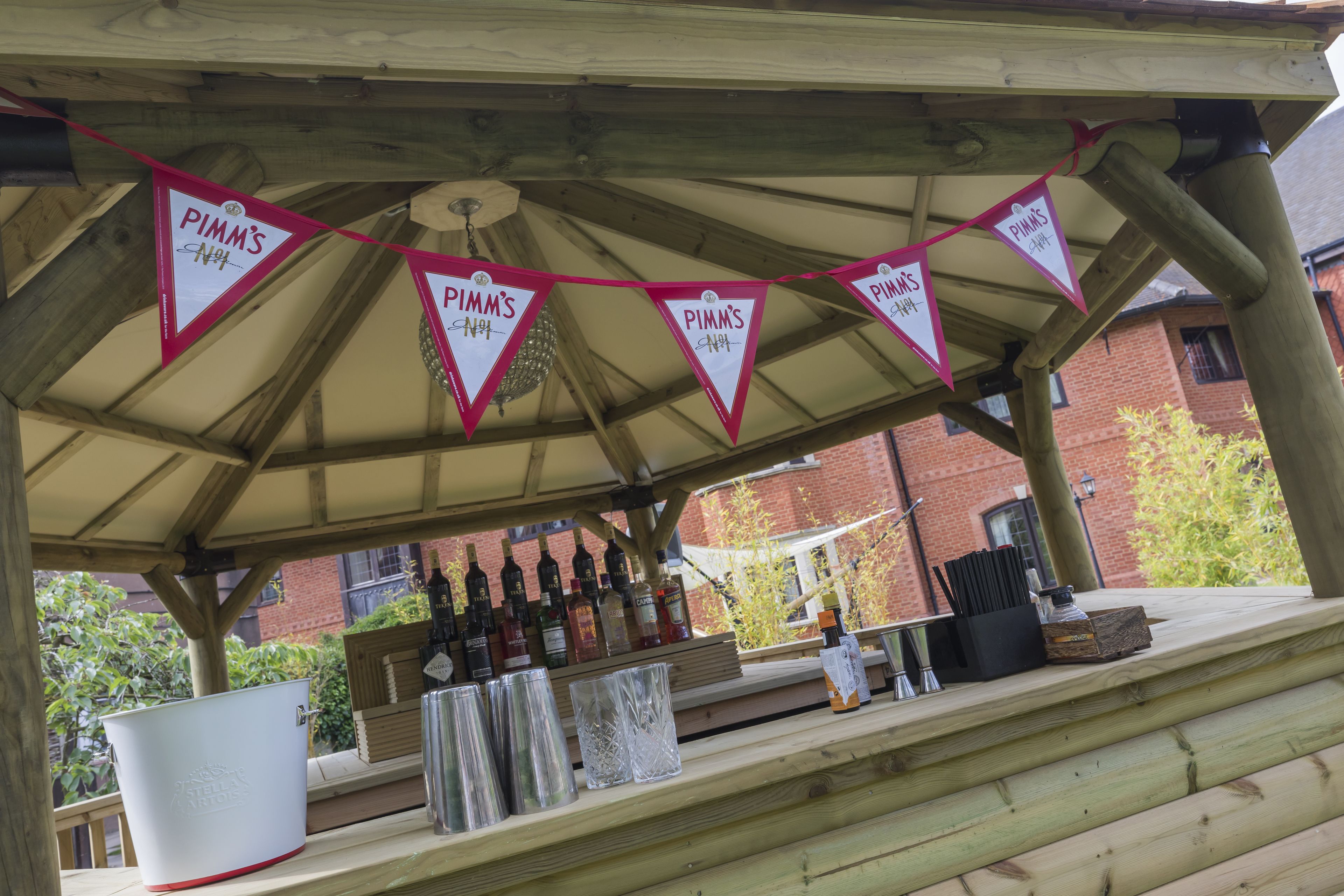 A covered outdoor bar area with a wooden structure, a row of bottles, and a white bucket.