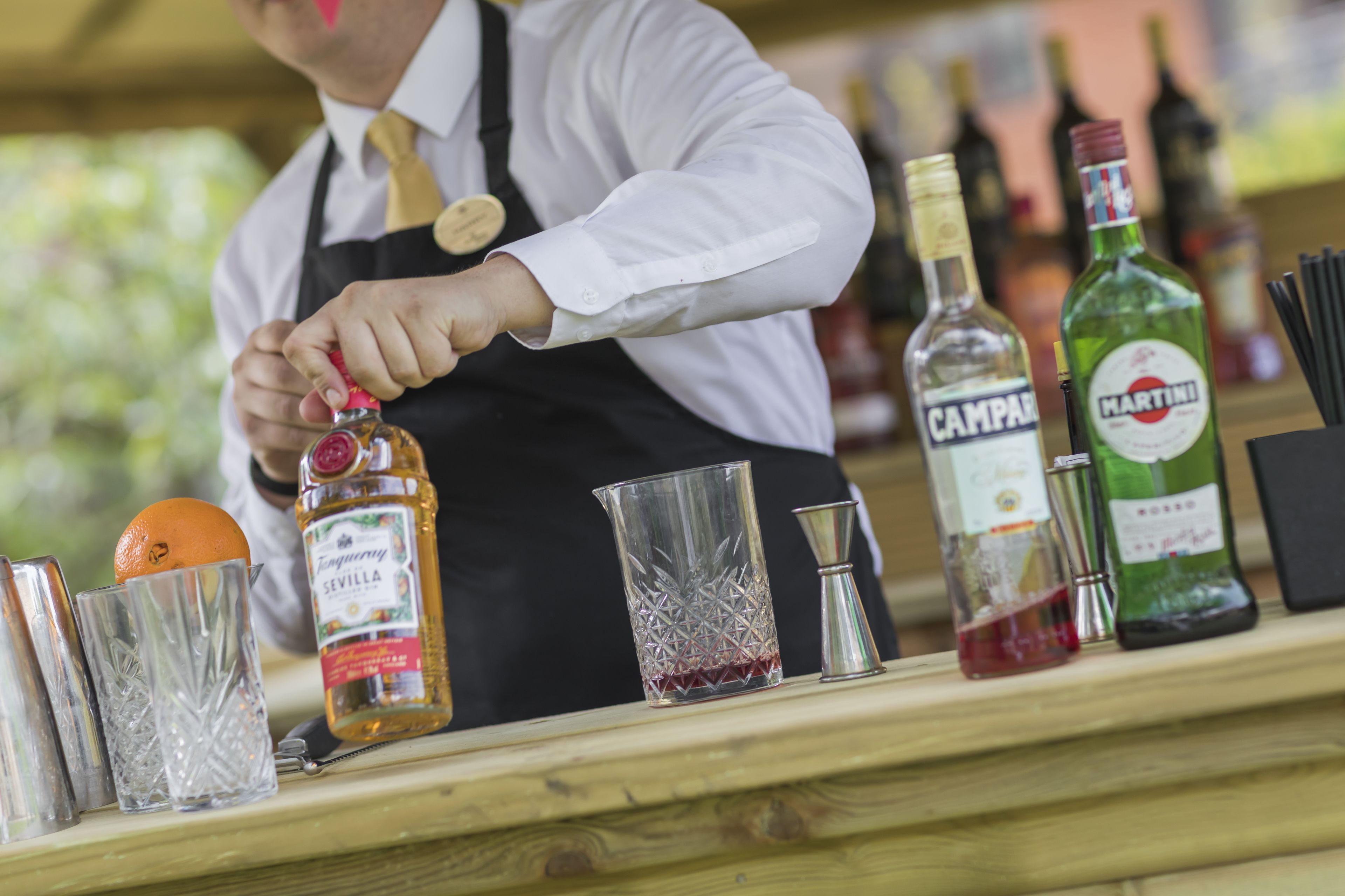 A bartender preparing a drink with a bottle of liquor, a glass pitcher, and various bar tools on a wooden counter.