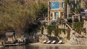 A stone building with a large window, a wooden deck with lounge chairs, and a small table by the water. - Kivotos Mykonos - Preferred Hotels & Resorts (Mykonos)