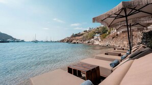 A beachside lounge with lounge chairs and a wooden table, overlooking a calm sea with boats and a rocky hillside in the background. - Kivotos Mykonos - Preferred Hotels & Resorts (Mykonos)