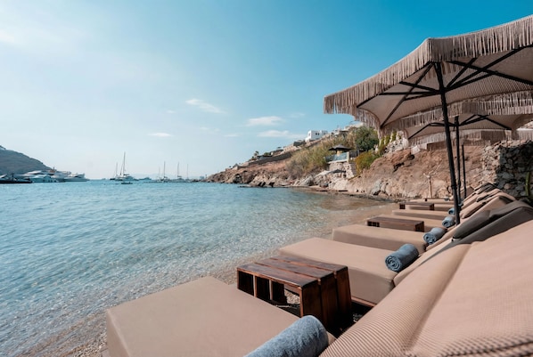 A beachside lounge with lounge chairs and a wooden table, overlooking a calm sea with boats and a rocky hillside in the background.
