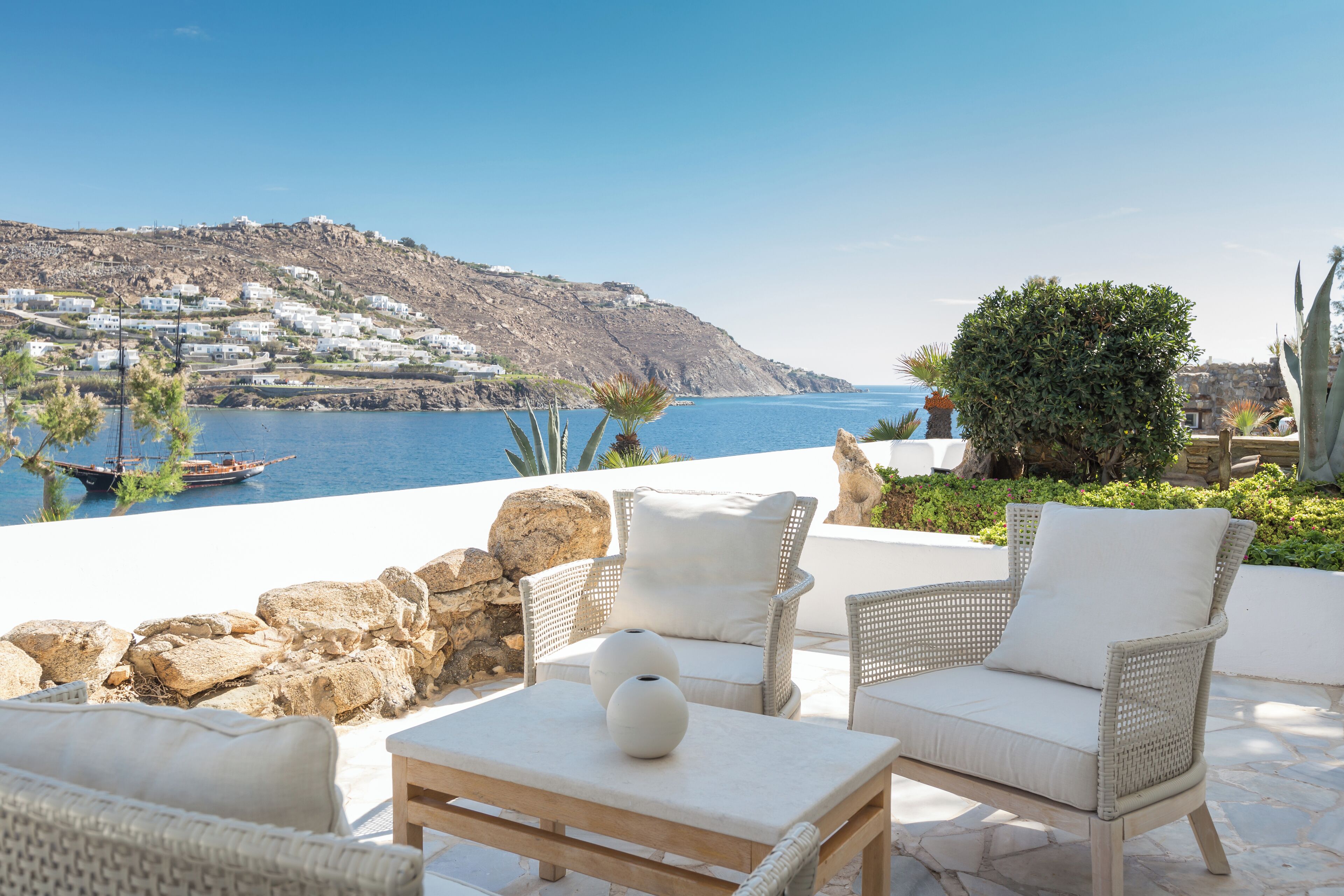 Eine Terrasse mit Korbmöbeln, einem Steintisch und Blick auf das Meer sowie ein Hügeldorf.