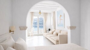 A minimalist bedroom with a large bed, a sofa, and a view of the sea through an arched doorway. - Kivotos Mykonos - Preferred Hotels & Resorts (Mykonos)