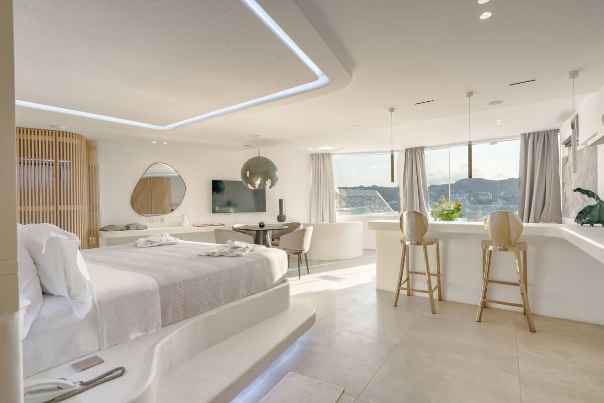Een moderne hotelkamer met een bed, een bureau, twee barkrukken, een groot raam met uitzicht op de stad en een ronde spiegel aan de muur.