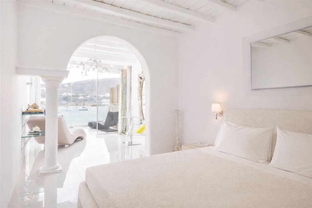 Een slaapkamer met witte muren, een groot bed, een nachtkastje, een lamp en uitzicht op de zee door een boogvormige deuropening.