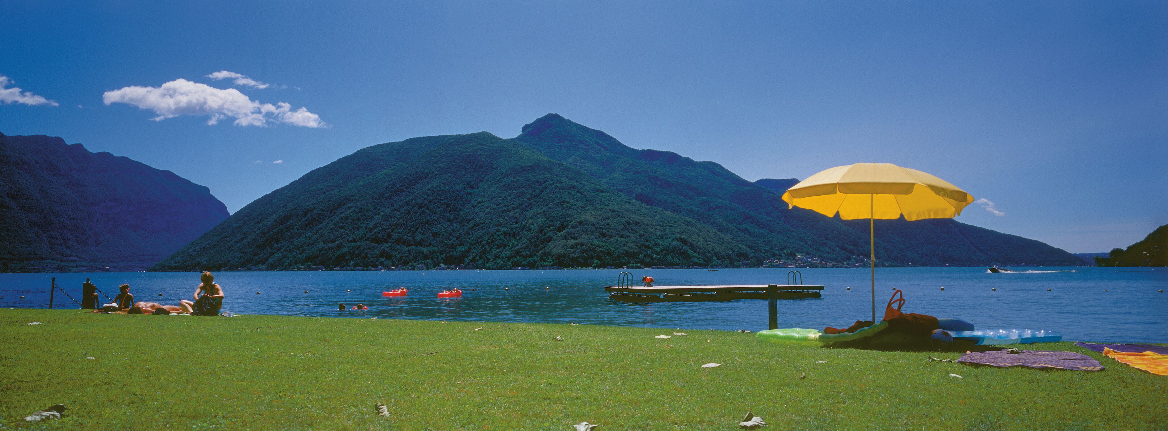 A lakeside scene with a grassy area, a dock, and a yellow umbrella.
