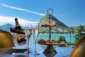 A table with a bottle of champagne, two glasses, and a dessert tray with strawberries and leaves, set against a scenic mountain view.