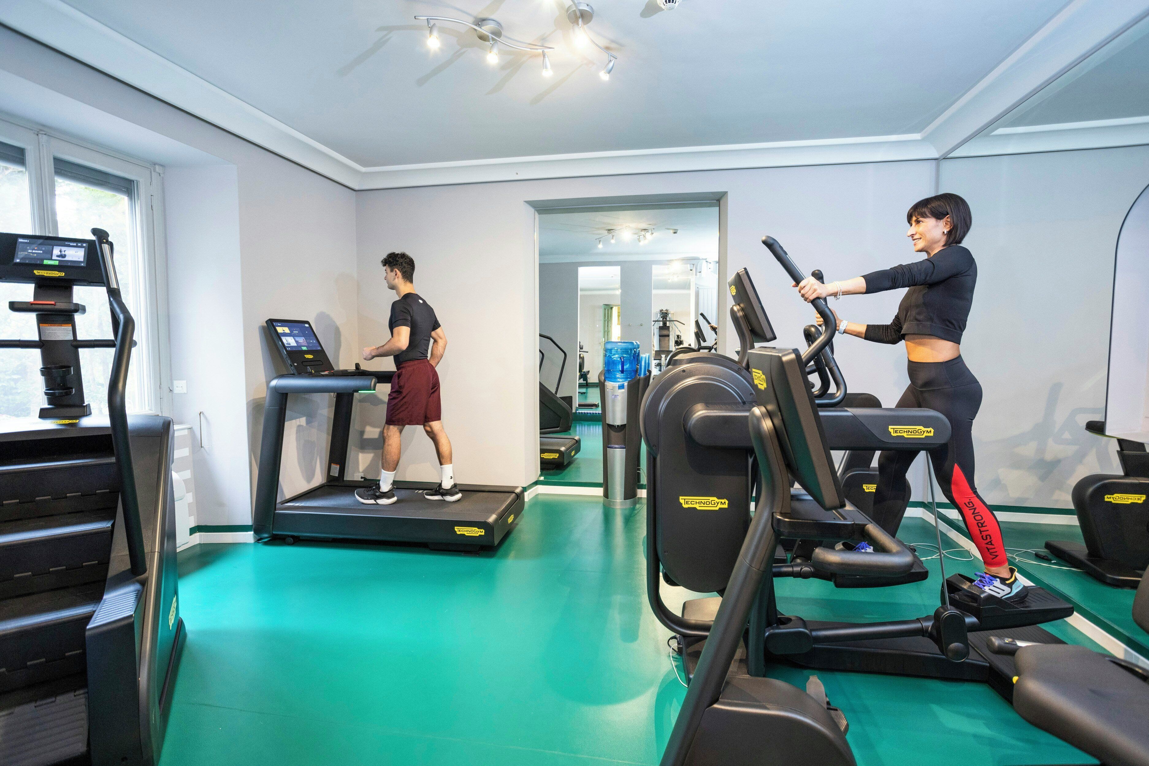Un gimnasio con cintas de correr, una persona caminando en una cinta y otra persona usando una bicicleta estática.