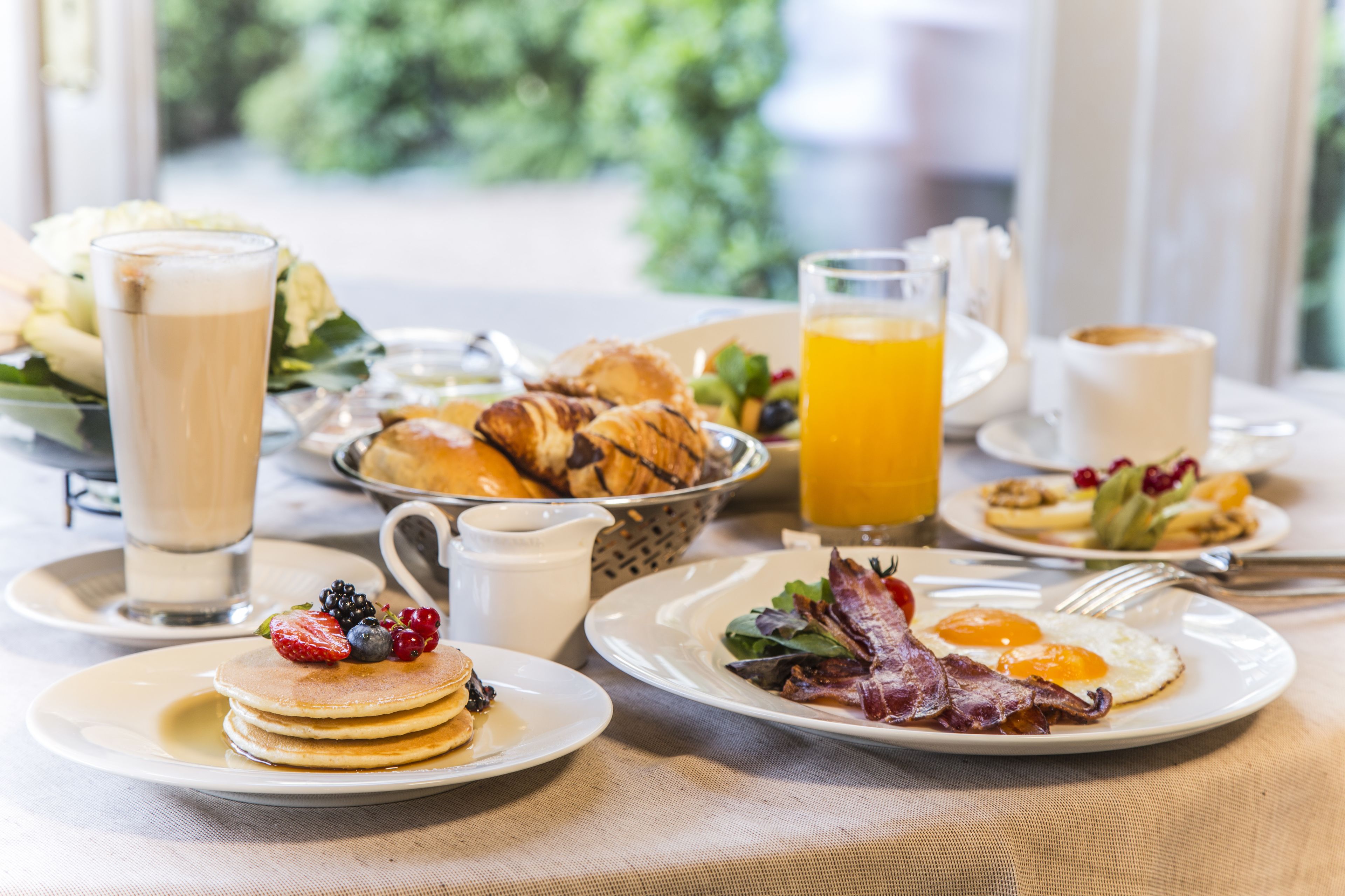 Desayuno con panqueques, huevos, tocino y fruta fresca en platos, junto con bebidas y pastelería.