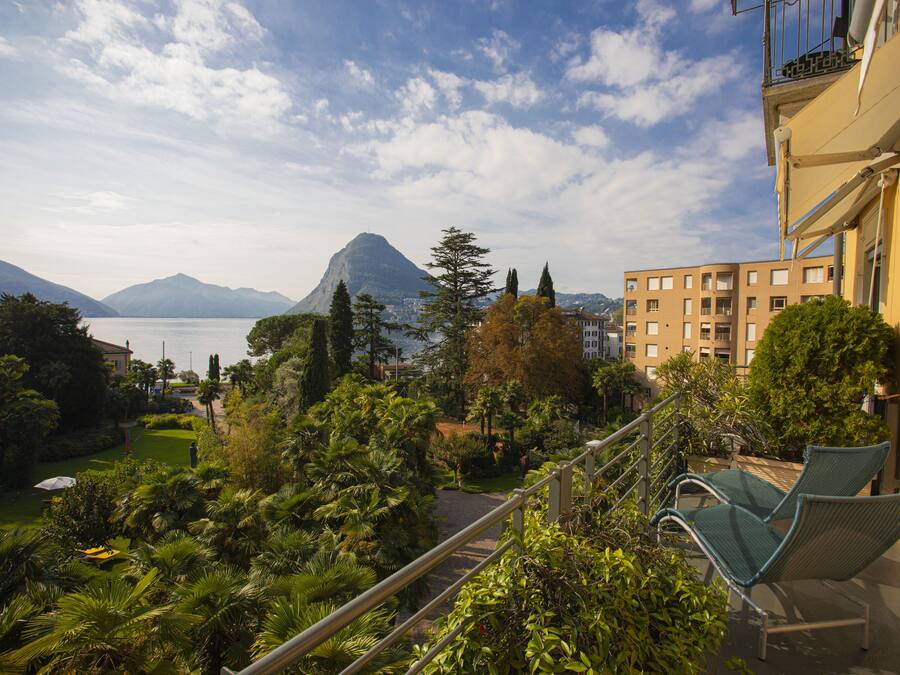 Un balcon avec deux fauteuils confortables donnant sur un jardin et un lac, avec des montagnes en toile de fond.