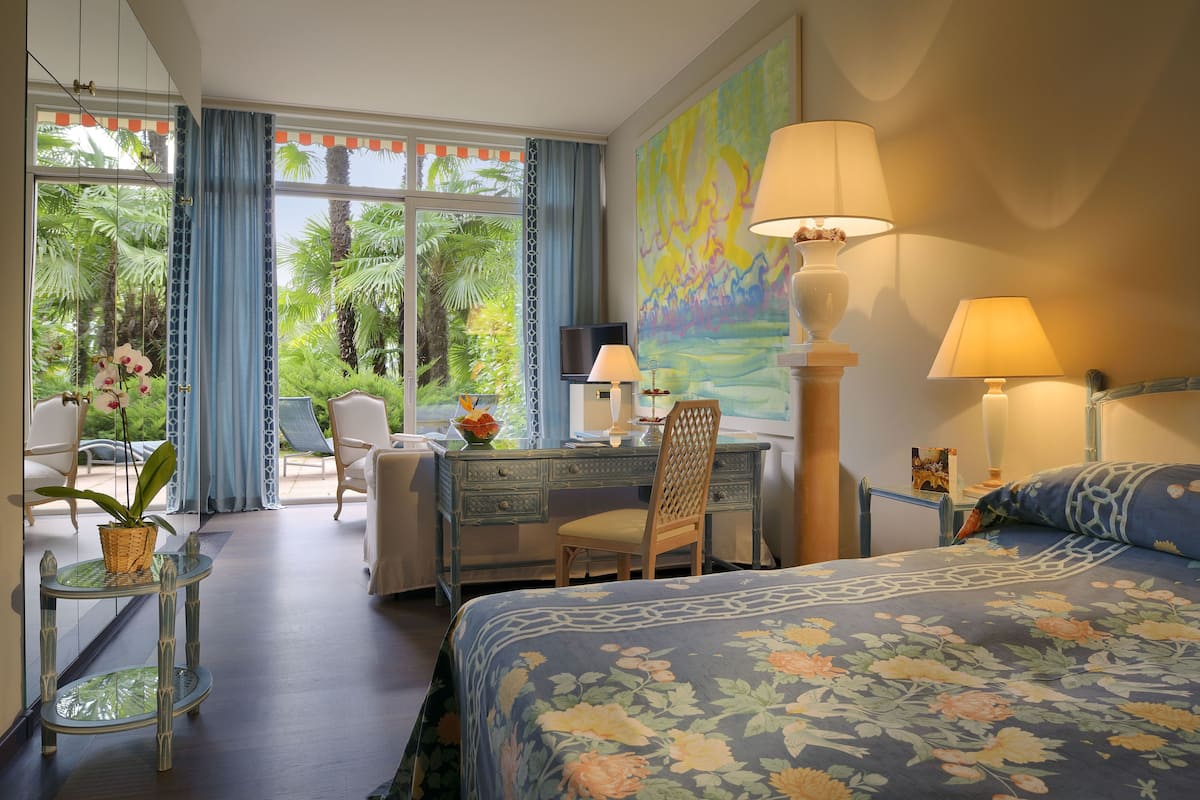 Habitación de hotel con cama, escritorio, sillas y vistas a palmeras.