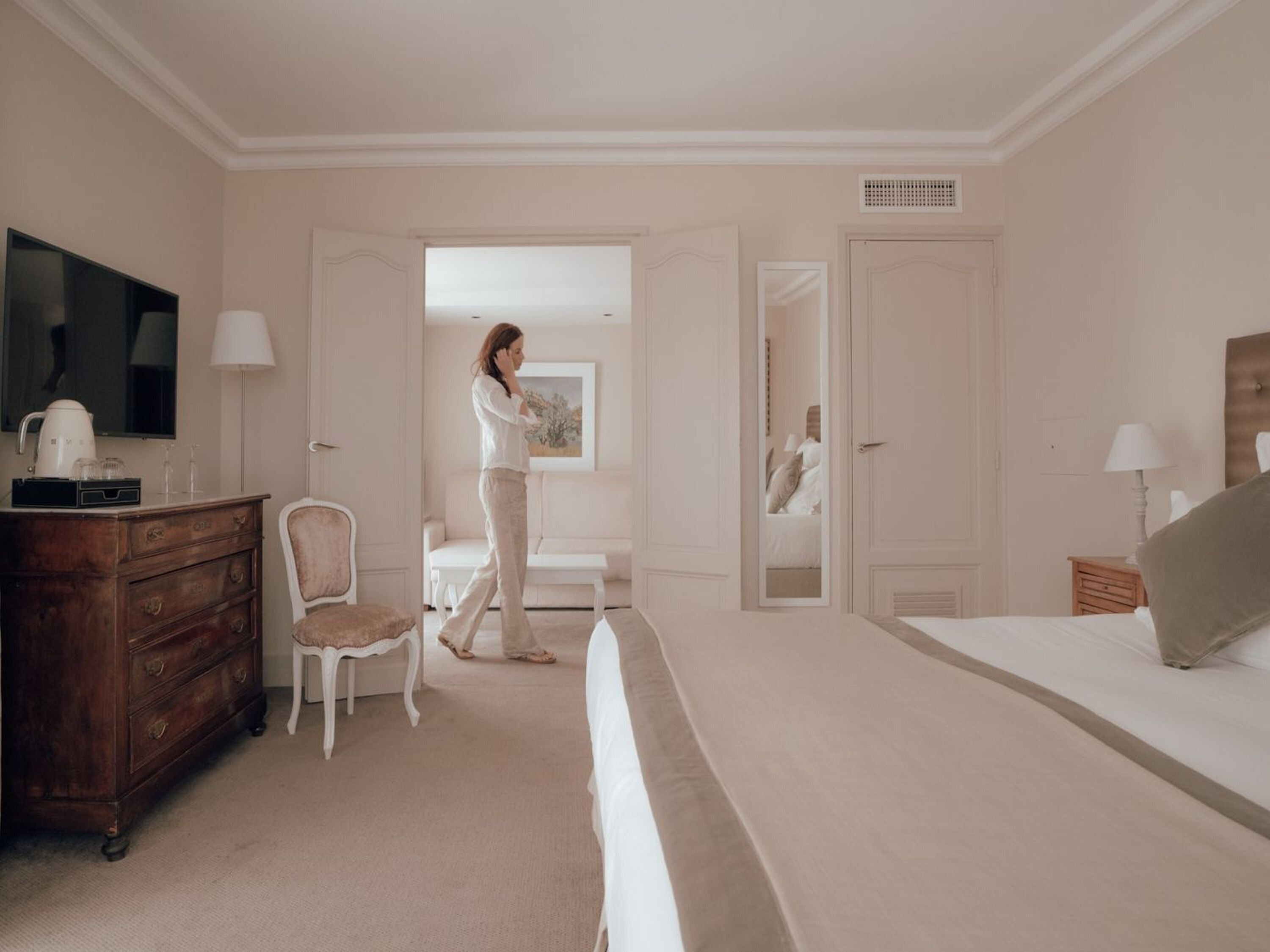 Ein Hotelzimmer mit einem Bett, einem Schminktisch, einem Sessel, einem Fernseher und einer Tür zu einem weiteren Raum.
