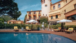 A pool area with lounge chairs and umbrellas, surrounded by a building with a tower and multiple balconies. - Ermitage de l'Oasis & Spa - Cannes Mandelieu (Mandelieu-La-Napoule)