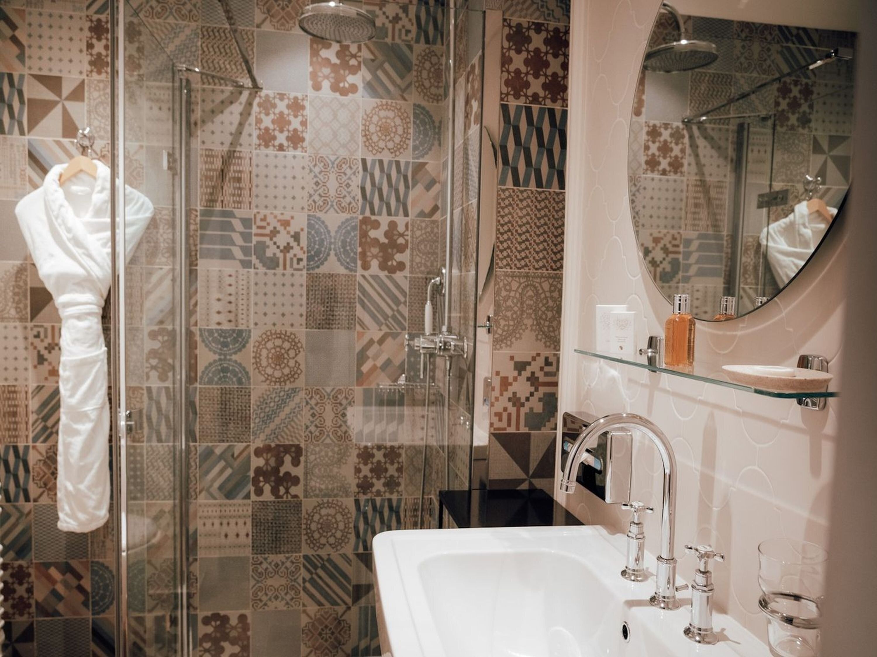 A bathroom with patterned tiles, a glass shower enclosure, a white sink, and a mirror with a round frame.