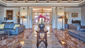 A spacious lobby with marble flooring, two sofas, a central table with a vase of flowers, and classical architectural elements like columns and a chandelier. - Ermitage de l'Oasis & Spa - Cannes Mandelieu (Mandelieu-La-Napoule)