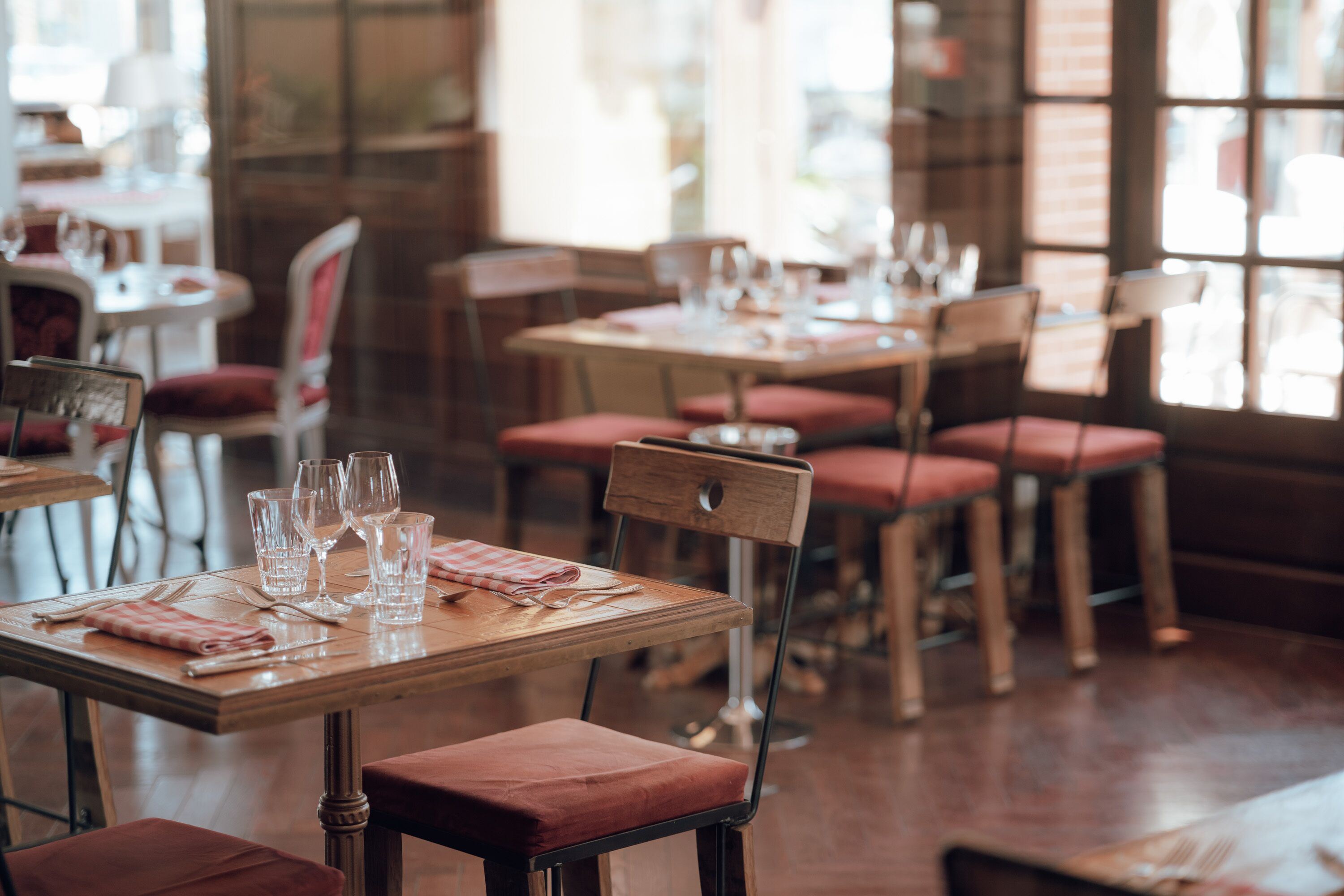 A restaurant interior with wooden tables and red cushioned chairs, some tables set with glasses and cutlery.