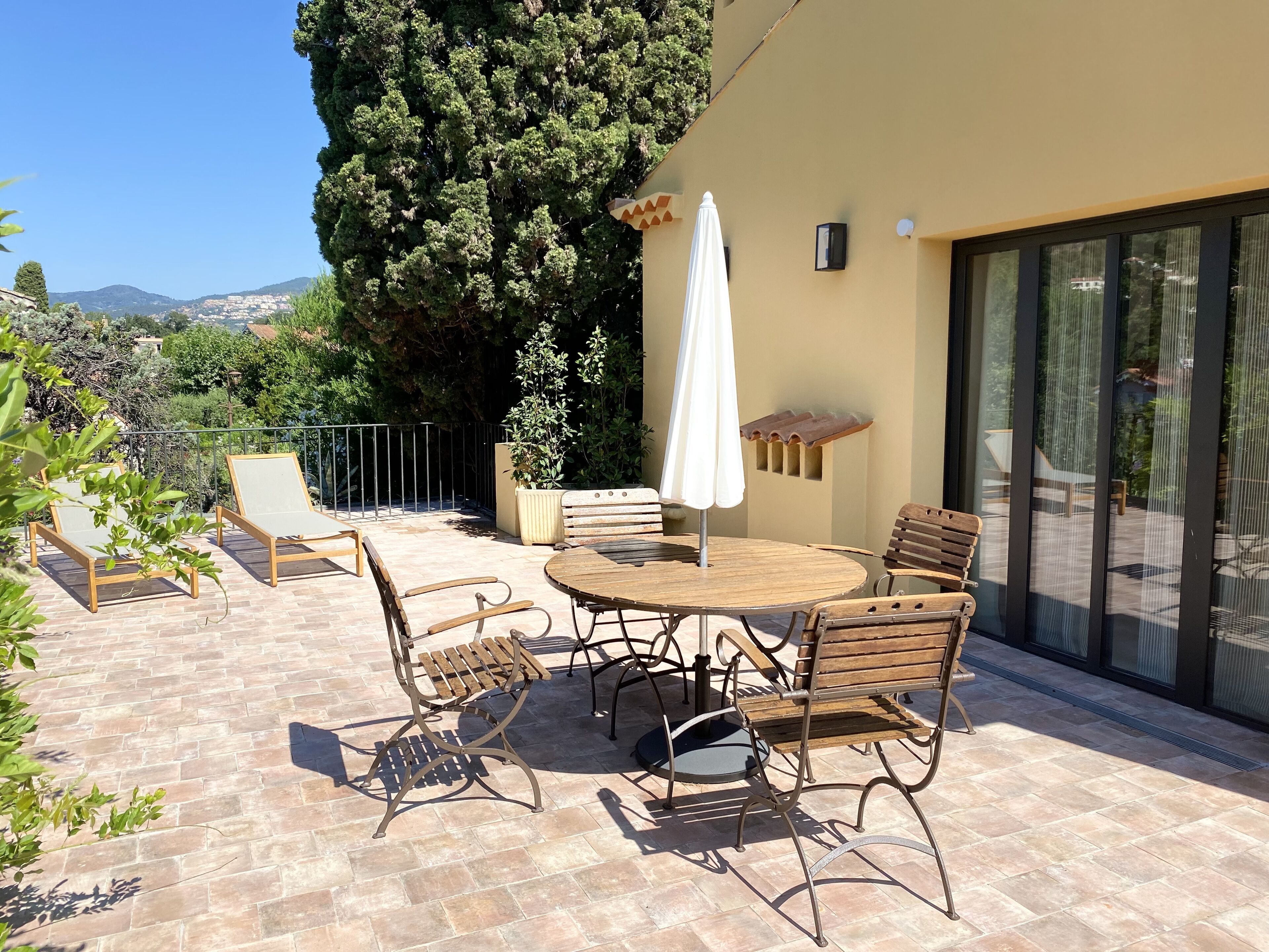 A patio with a round table and chairs, a white umbrella, and a view of the surrounding landscape.