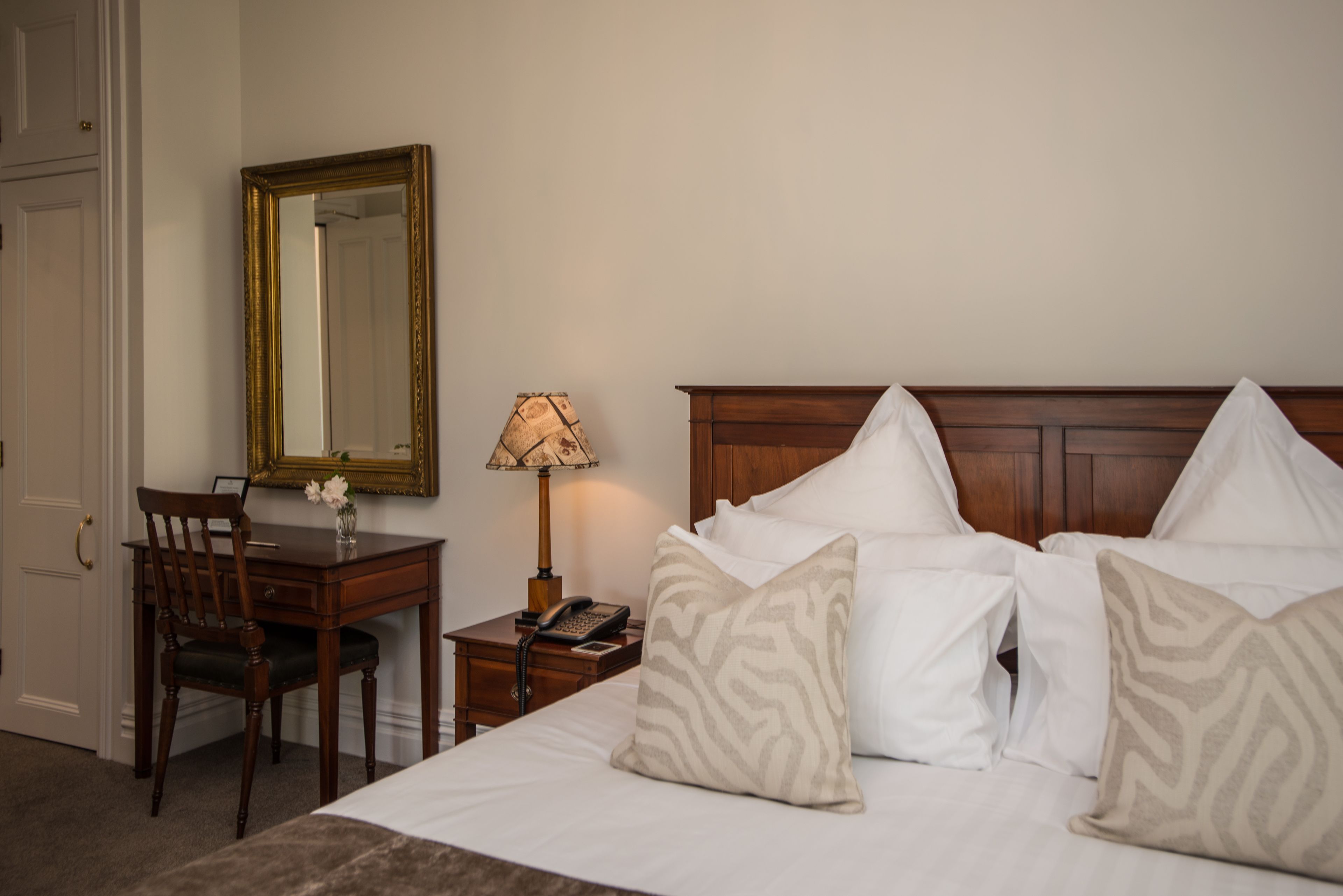 A hotel room with a wooden bed, white pillows, a wooden nightstand with a lamp, a wooden chair, and a mirror on the wall.