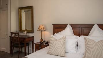 A hotel room with a wooden bed, white pillows, a wooden nightstand with a lamp, a wooden chair, and a mirror on the wall.