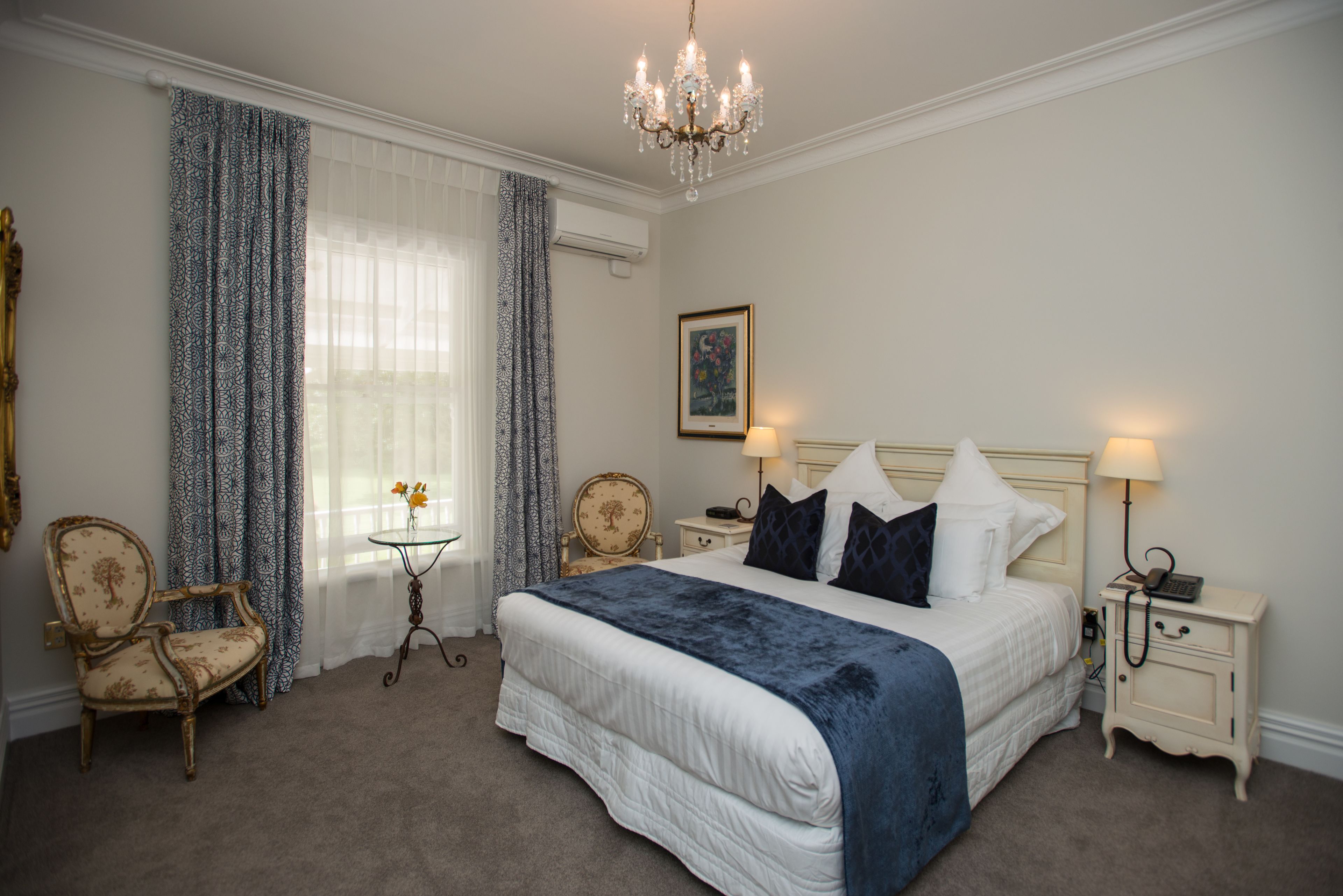 A bedroom with a large bed, two bedside tables, a chair, a small table with flowers, and a chandelier.
