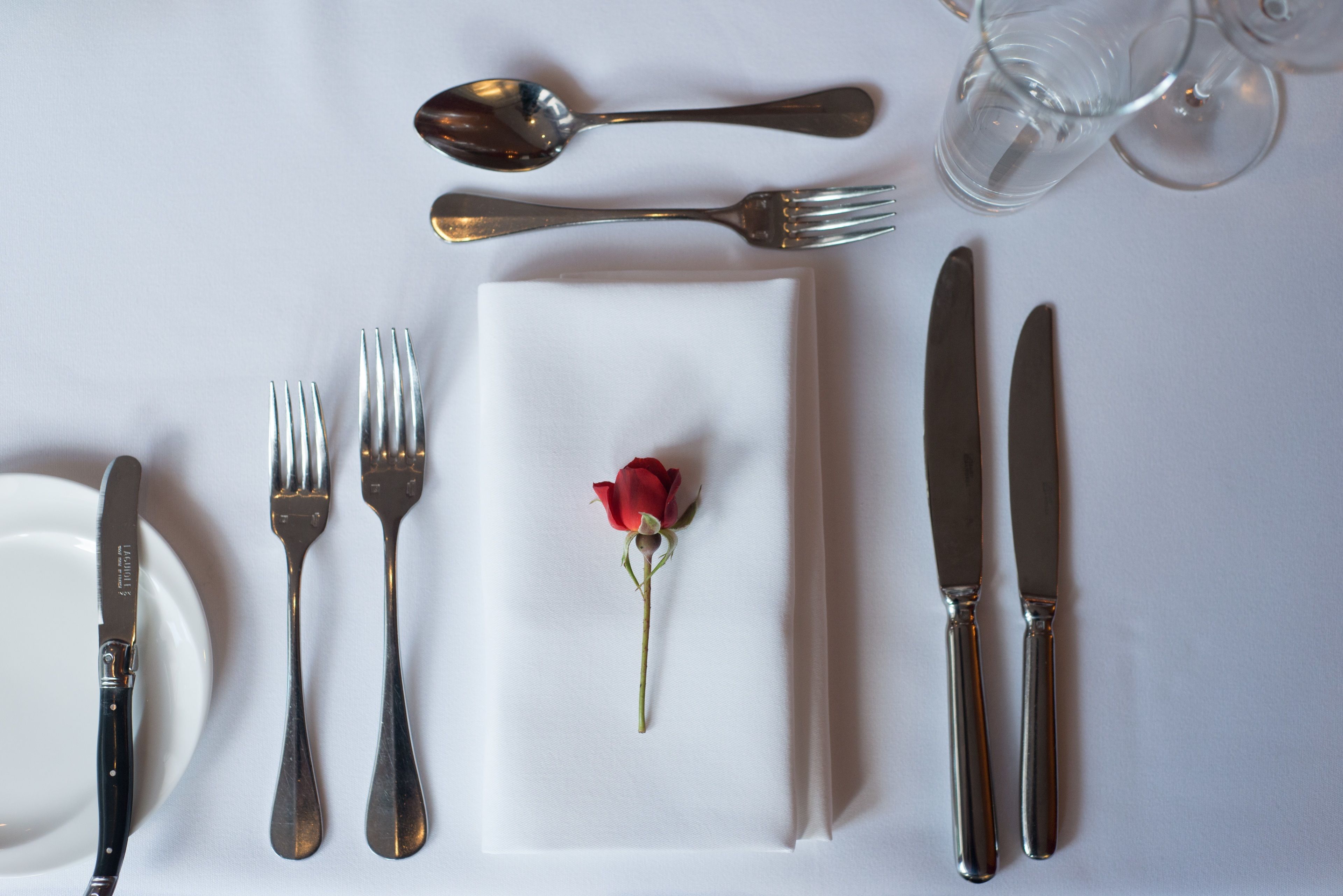 A table setting with a white napkin, a red rose, and silverware including a knife, fork, and spoon.