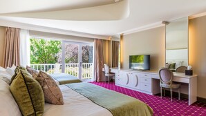 Habitación de hotel con cama, escritorio, silla, televisor y ventana grande con cortinas.