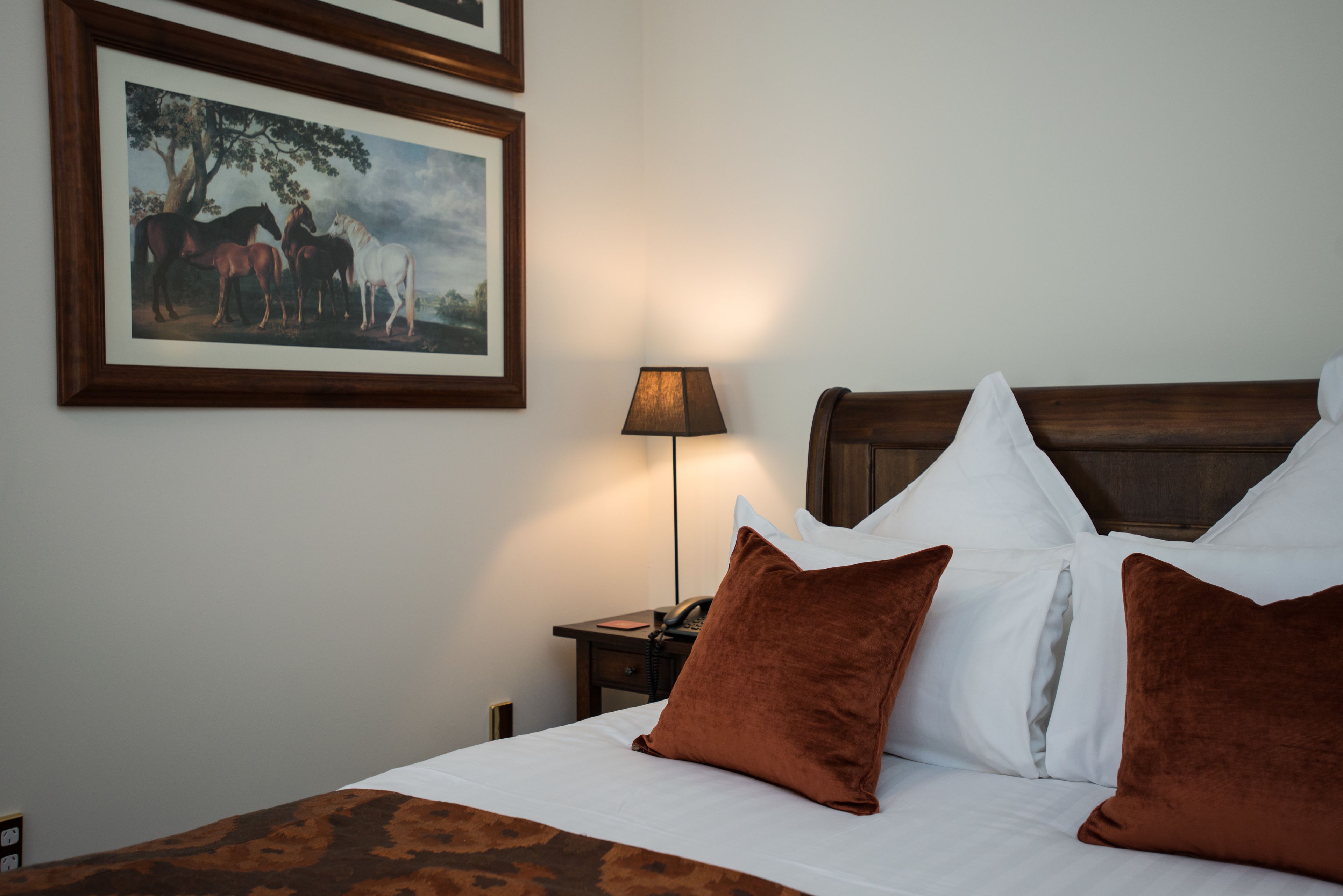 A neatly made bed with white linens and brown pillows, a wooden headboard, a nightstand with a lamp, and a framed painting of horses on the wall.