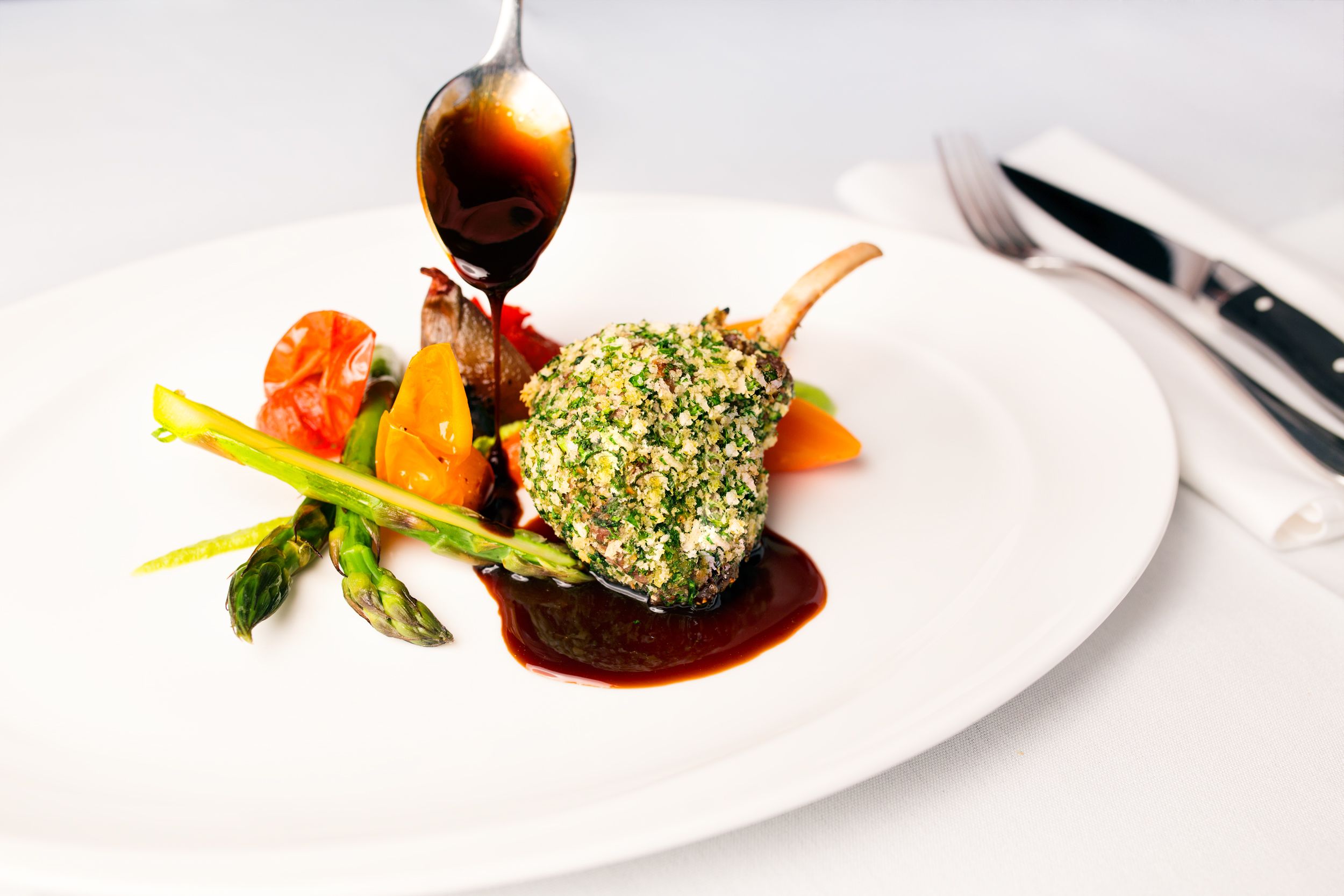Un plato blanco con un palillo de cordero, espárragos y verduras, además de una cuchara con salsa.